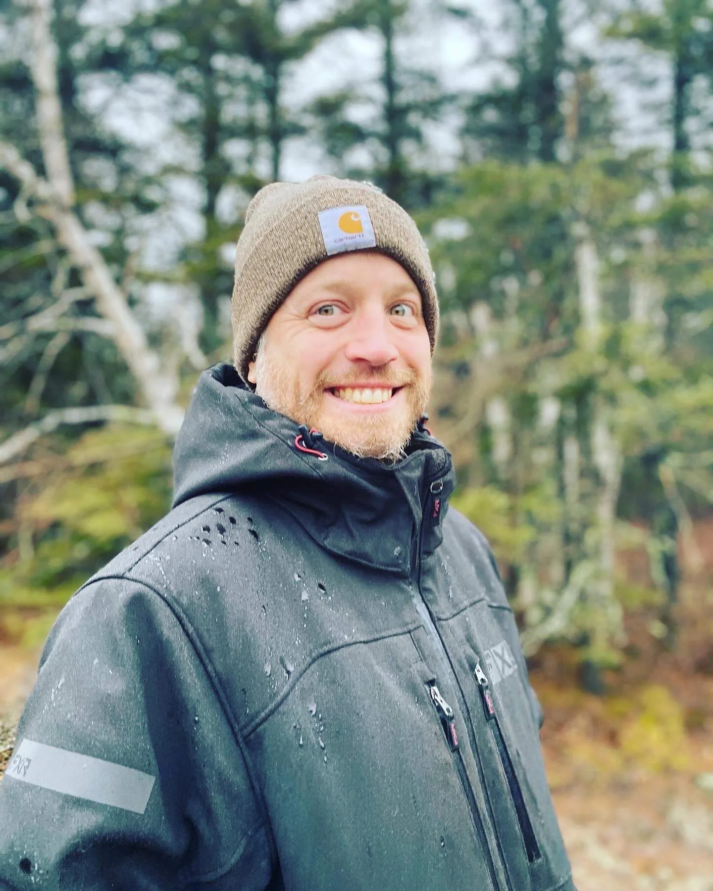A smiling man in a black jacket and gray beanie in a wooded outdoor setting.