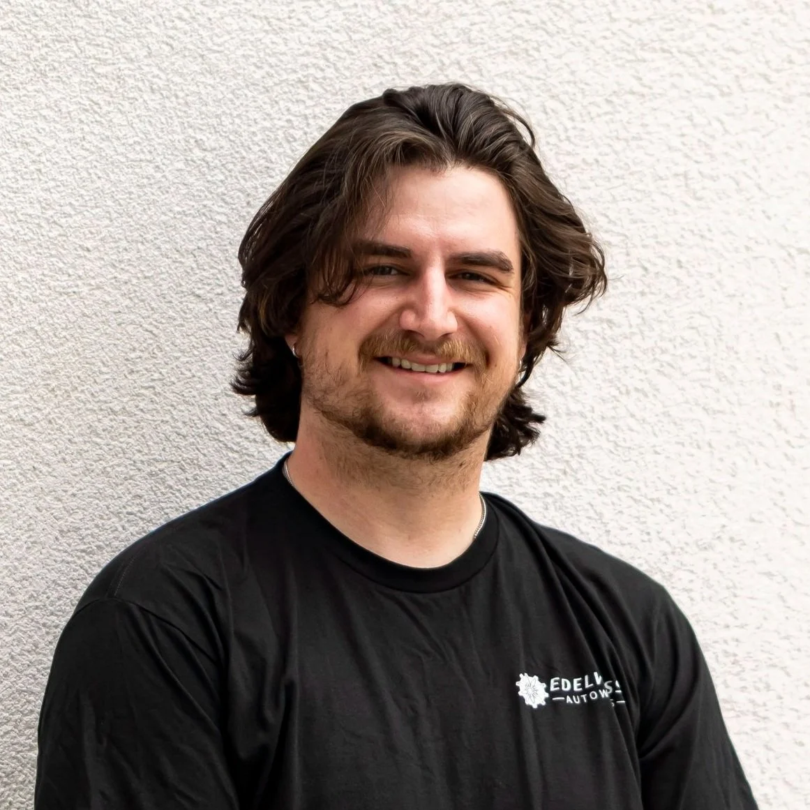 A smiling young man with long, wavy brown hair and a beard, wearing a black t-shirt with a logo, standing against a textured light-colored wall.
