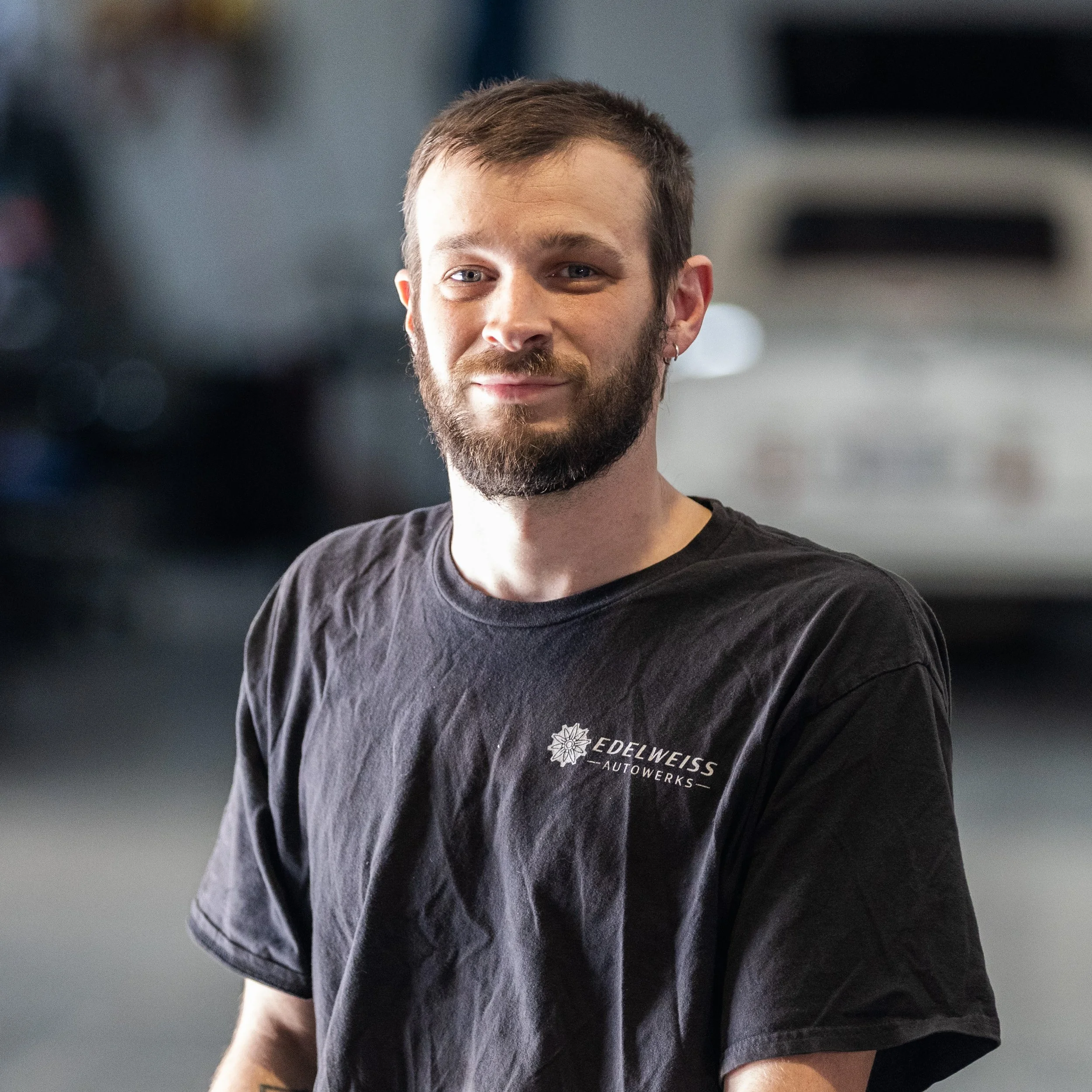 A man with a beard wearing a black T-shirt with a small logo, standing in front of a textured white wall.