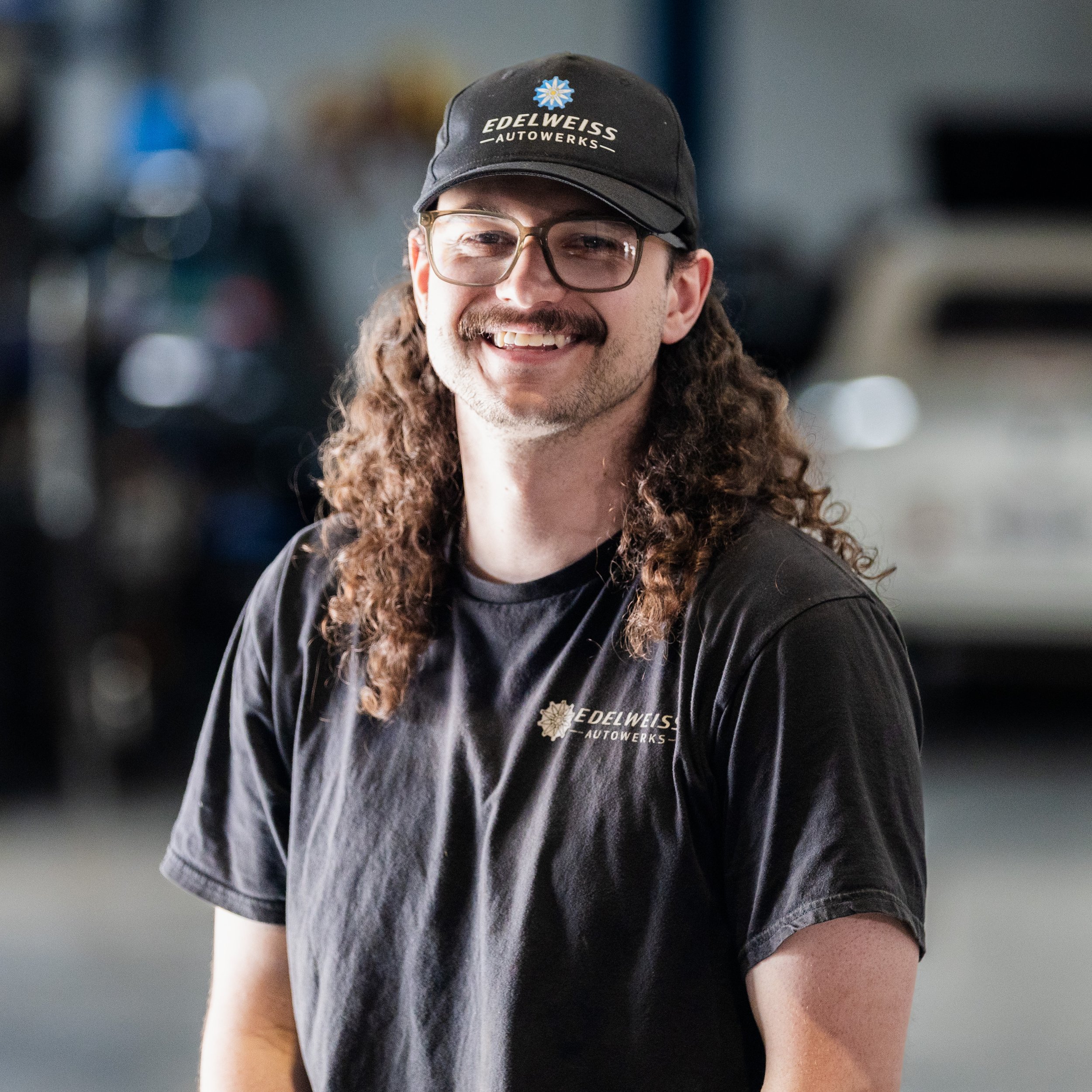 A smiling man with curly hair, wearing glasses, a black t-shirt with Edelweiss AutoWorks logo, and a Snap-on Tools cap, standing against a white textured wall.