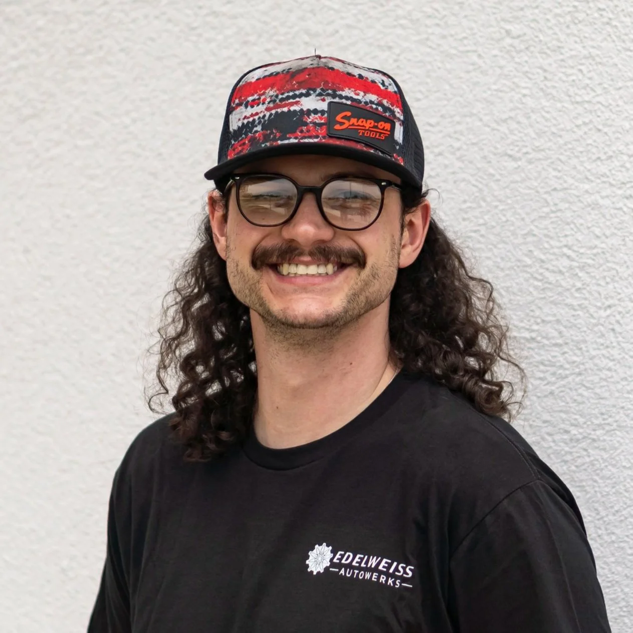 A smiling man with curly hair, wearing glasses, a black t-shirt with Edelweiss AutoWorks logo, and a Snap-on Tools cap, standing against a white textured wall.