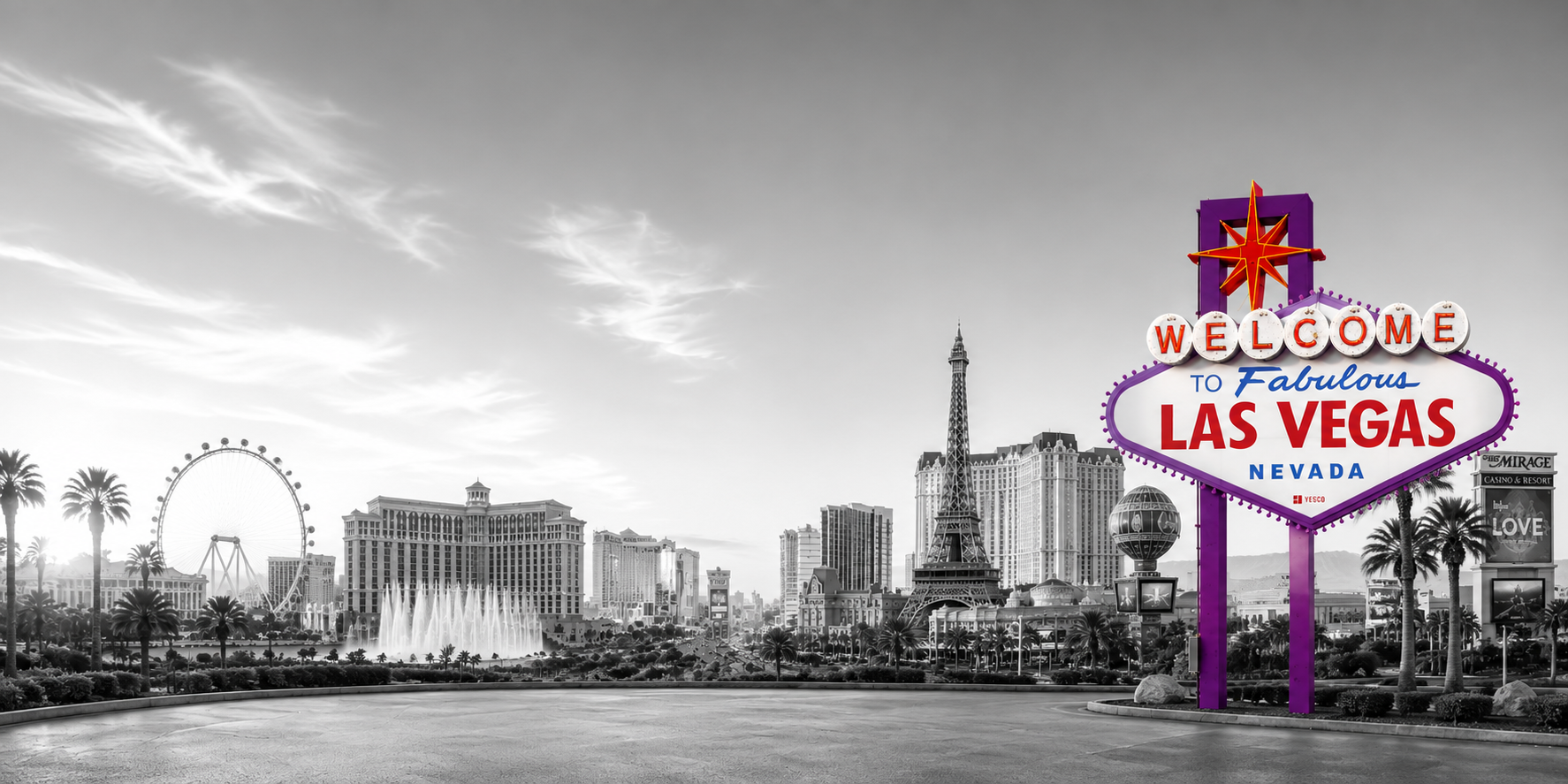 Black and white view of the Las Vegas skyline with a colorful welcome sign in the foreground that reads "Welcome to Fabulous Las Vegas, Nevada." The skyline features a Ferris wheel, the Las Vegas Eiffel Tower replica, and other tall buildings, with palm trees lining the street.