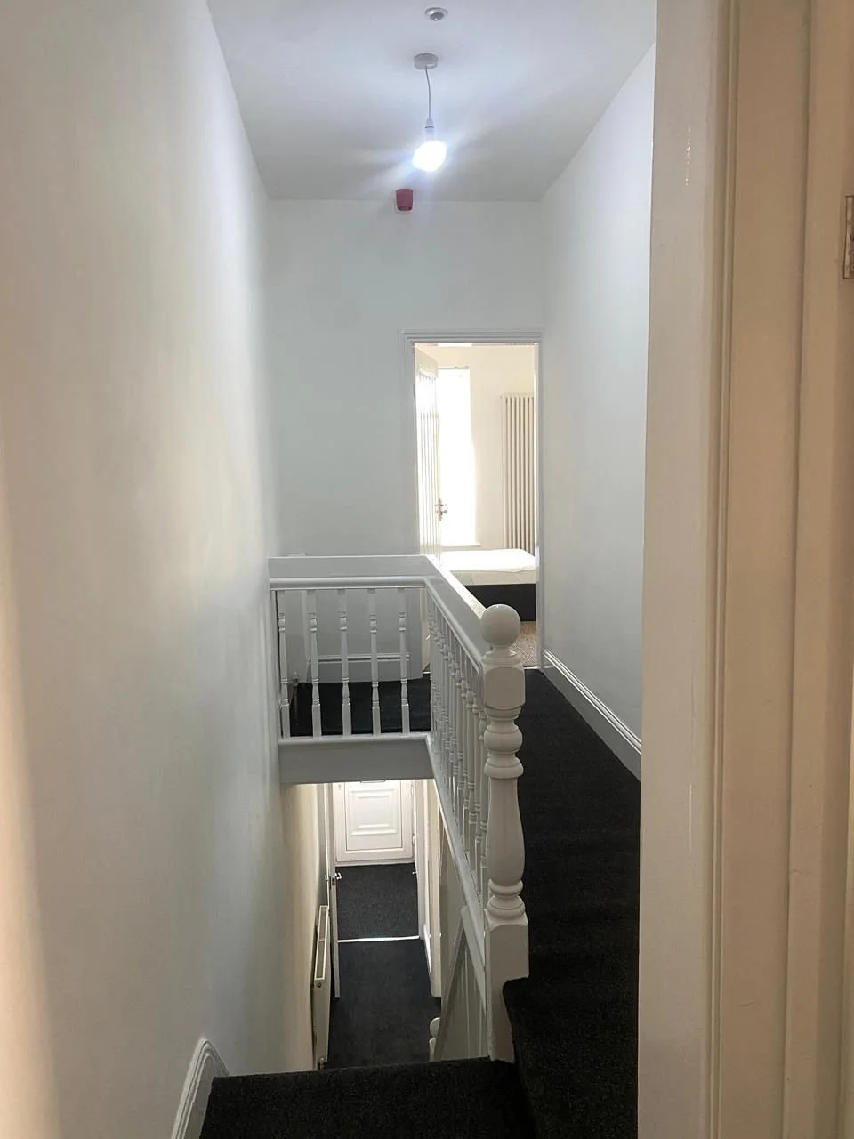 View of a hallway in a house with white walls, black carpeted floors, a staircase with white railings, and a door at the end of the hallway leading to a room with light coming in.