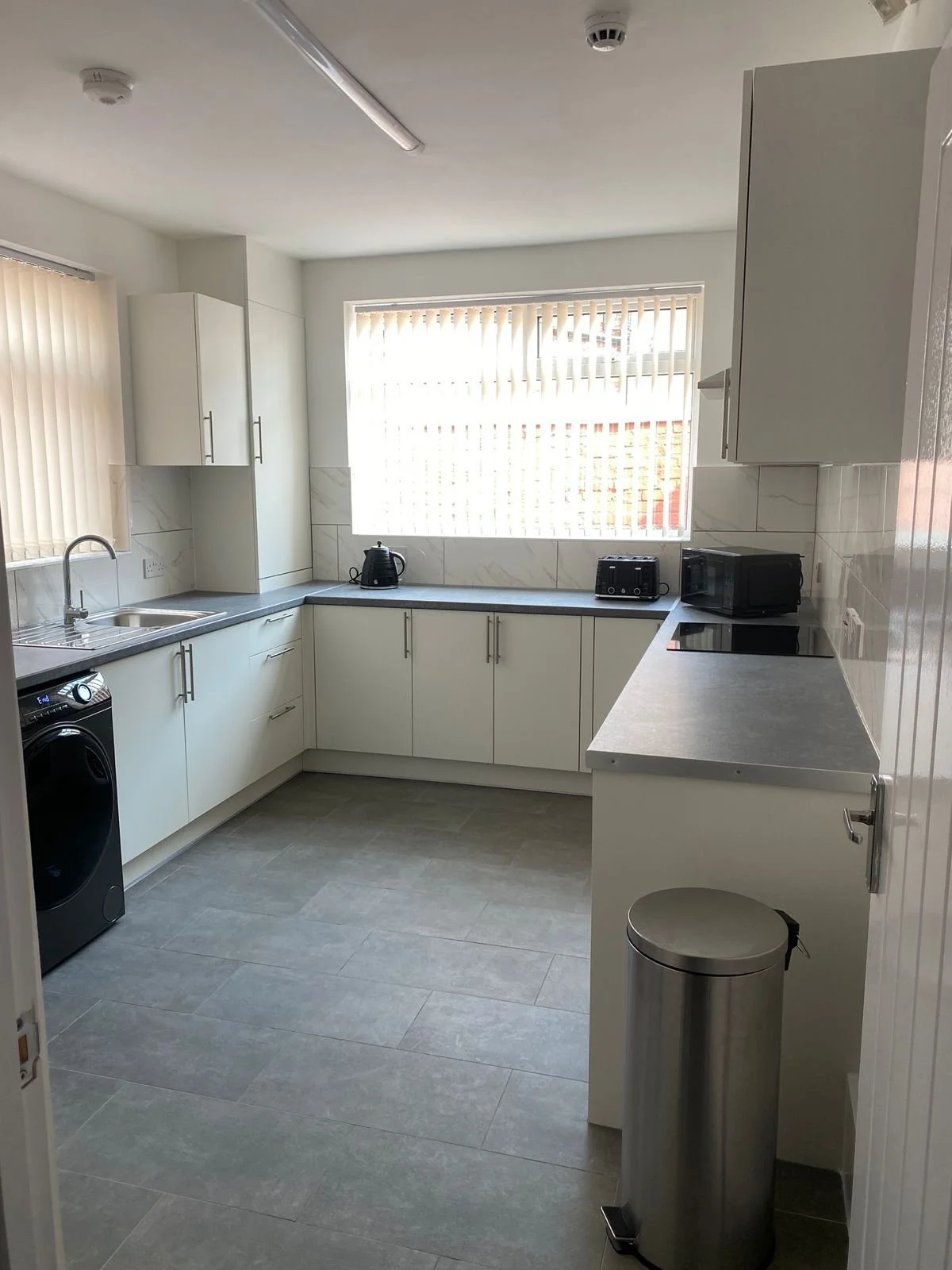 Modern kitchen with white cabinets, gray countertops, a stainless steel trash bin, and appliances including a washing machine, toaster, kettle, and microwave. Large window with vertical blinds.