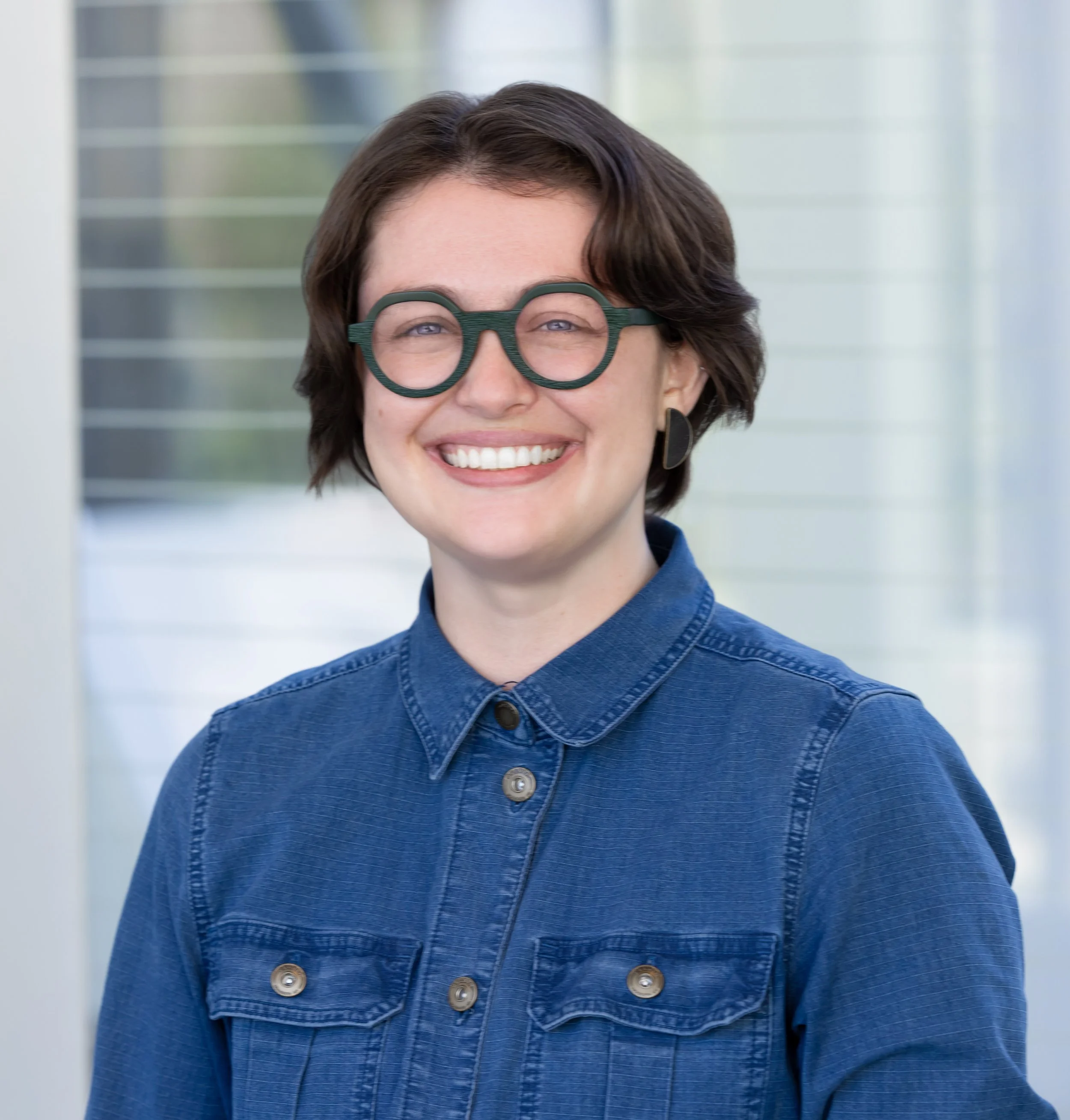 A woman with short brown hair, wearing round green glasses, a blue denim shirt, and black earrings, smiling indoors.