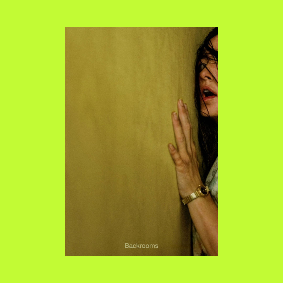 A woman with dark hair and glasses, wearing a watch, presses her hand against a yellow wall with a surprised or distressed expression.