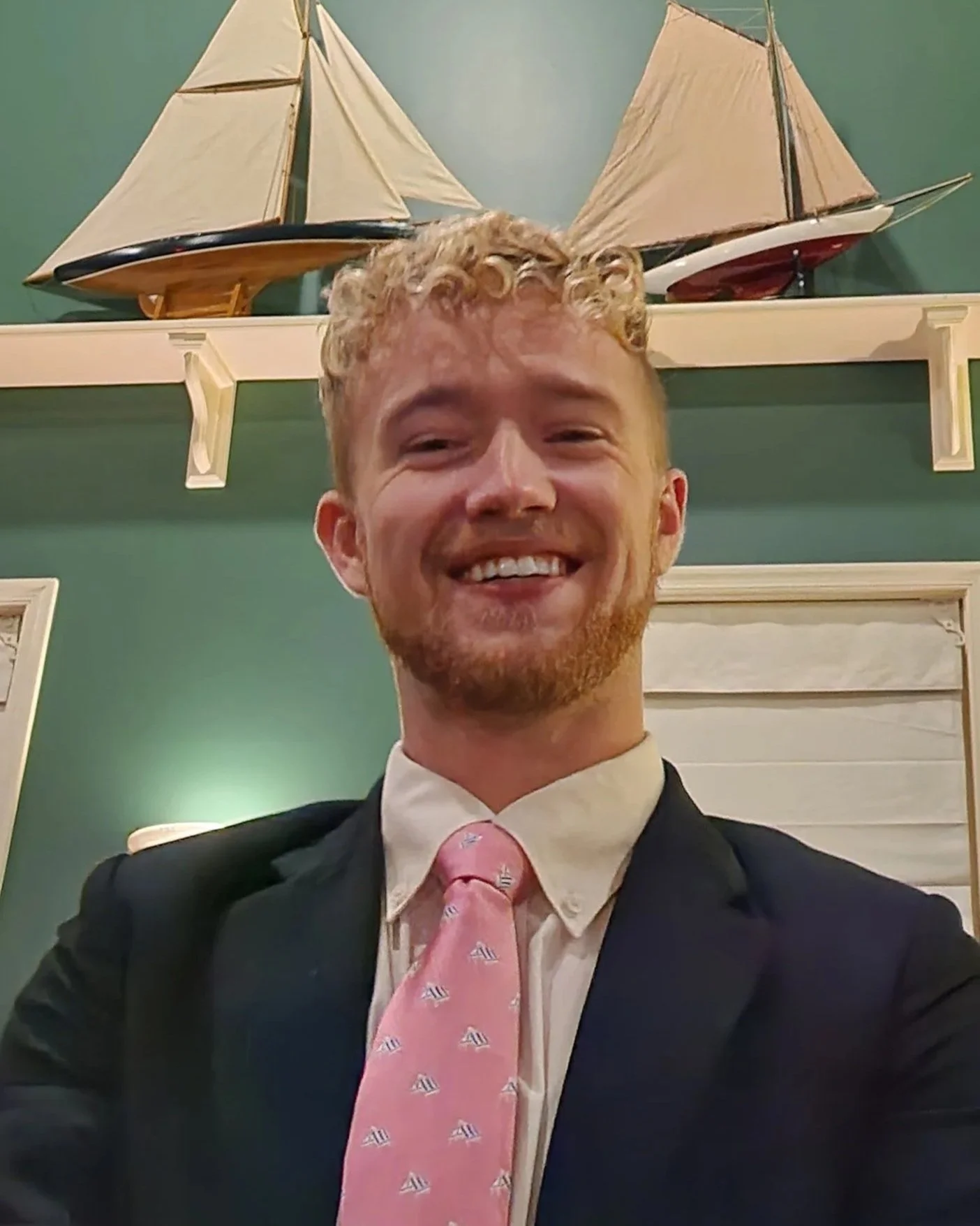 A smiling young man in a suit and pink tie taking a selfie in front of a green wall with two model sailboats displayed above him.
