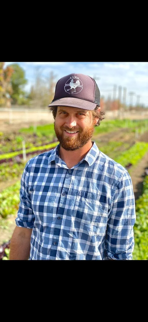 A young man with a beard, plaid shirt, and black baseball cap, standing in front of a small farm.
