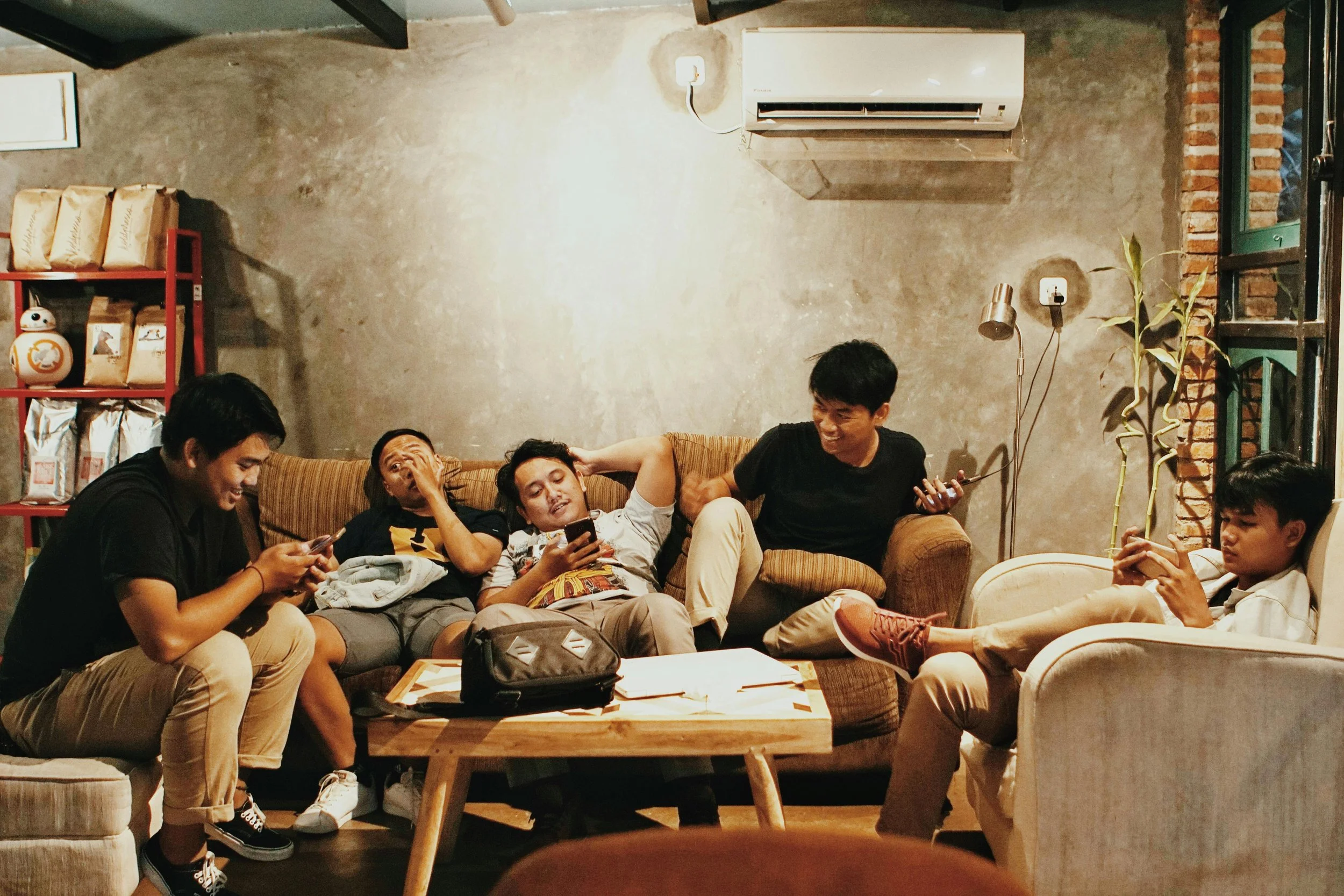 A group of Southeast Asian young men sit in a cozy café lounge, laughing, chatting, and checking their phones. The atmosphere feels warm, casual, and communal.