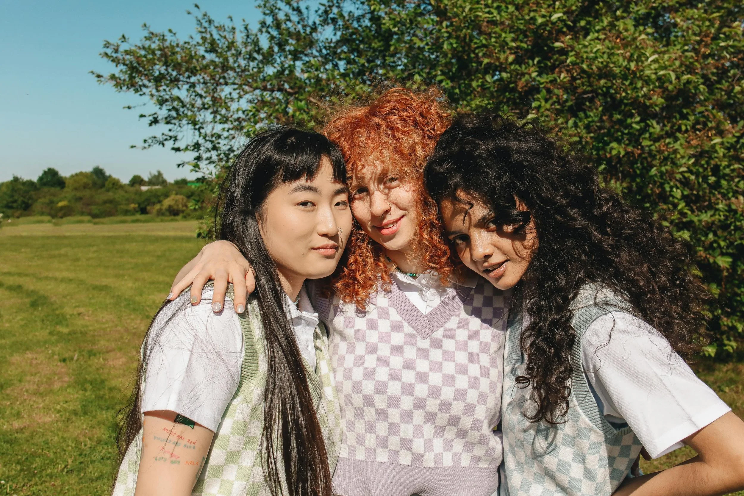 Three women of color stand closely together outdoors, wearing checkered vests and smiling gently in the sunlight.