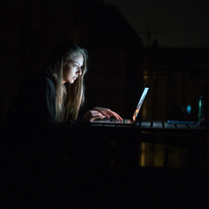 A woman works intensely on her laptop in a dark room, illuminated by the screen’s glow.