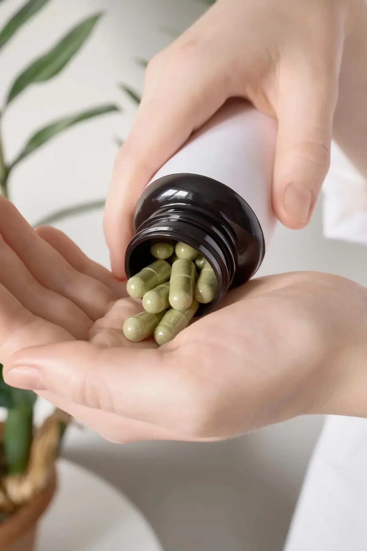 A woman's hand pours supplement capsules from a bottle into her palm, with a plant blurred in the background.