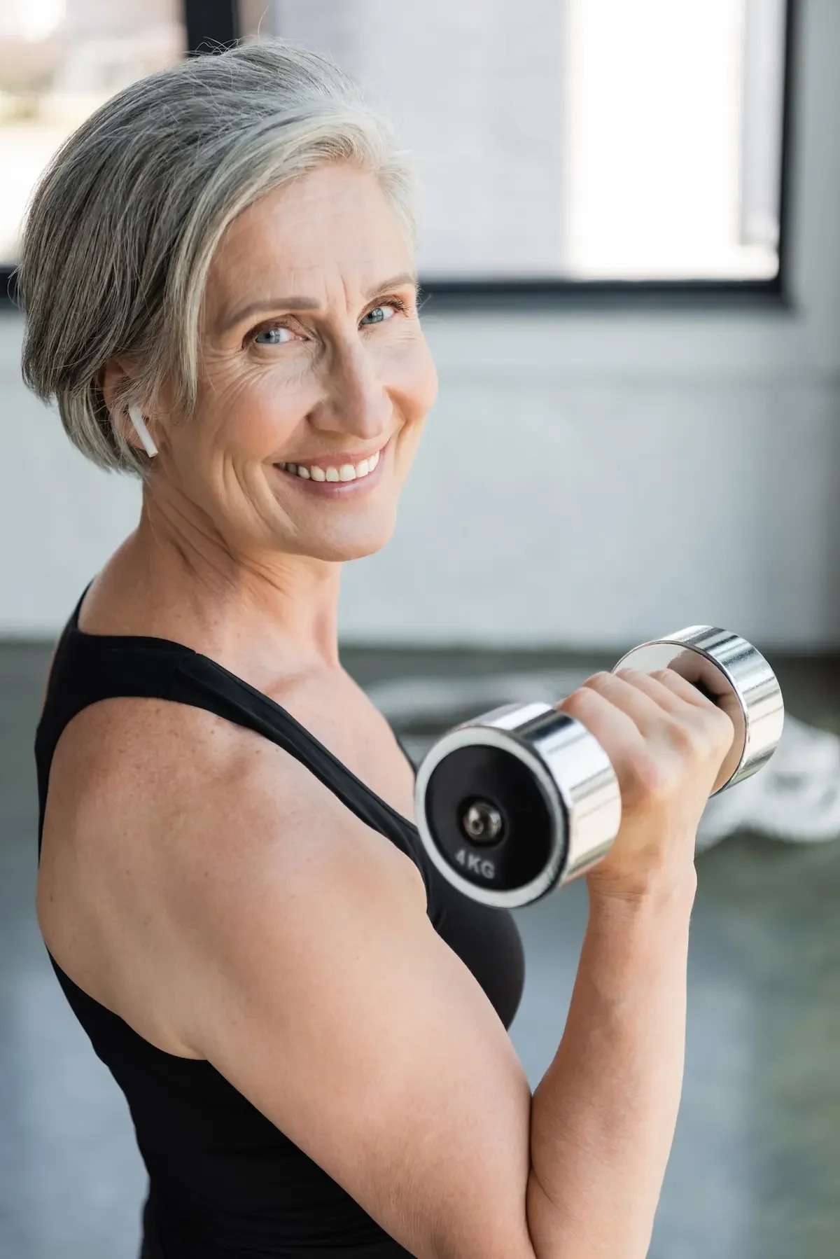 Midlife woman in athletic wear lifts hand weights, smiling at the camera; she has short gray hair and is wearing wireless earbuds.