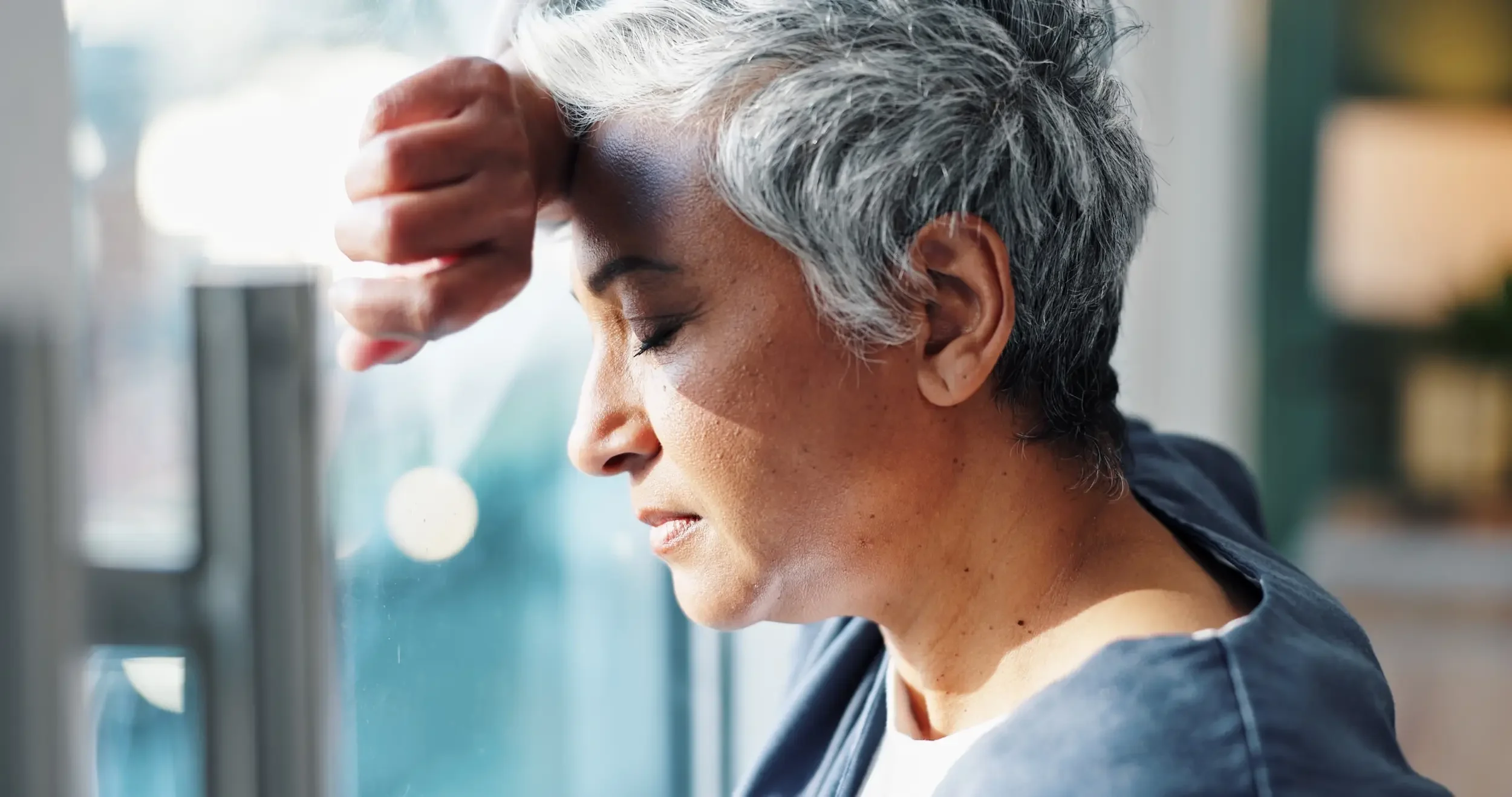Older woman with short gray hair leans against a window, eyes closed and hand on forehead, appearing tired or stressed, possibly experiencing uncomfortable sweating.