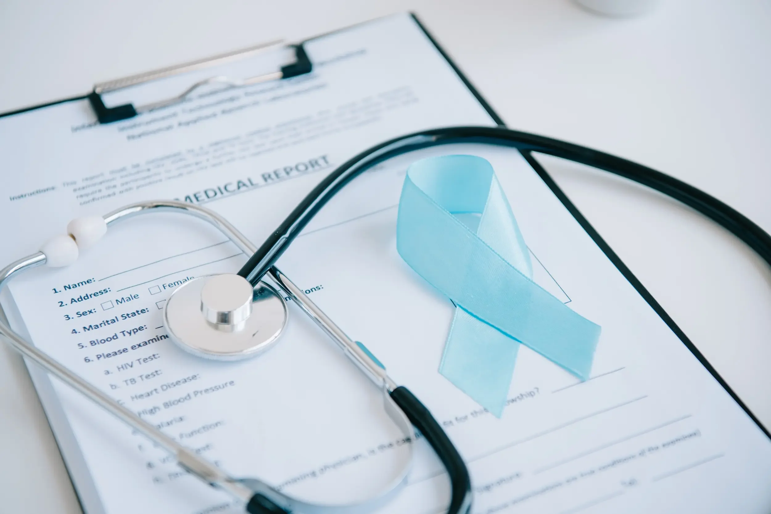 A stethoscope and a blue awareness ribbon rest on a medical report form attached to a clipboard, symbolizing the importance of generic cancer screening in preventive healthcare.
