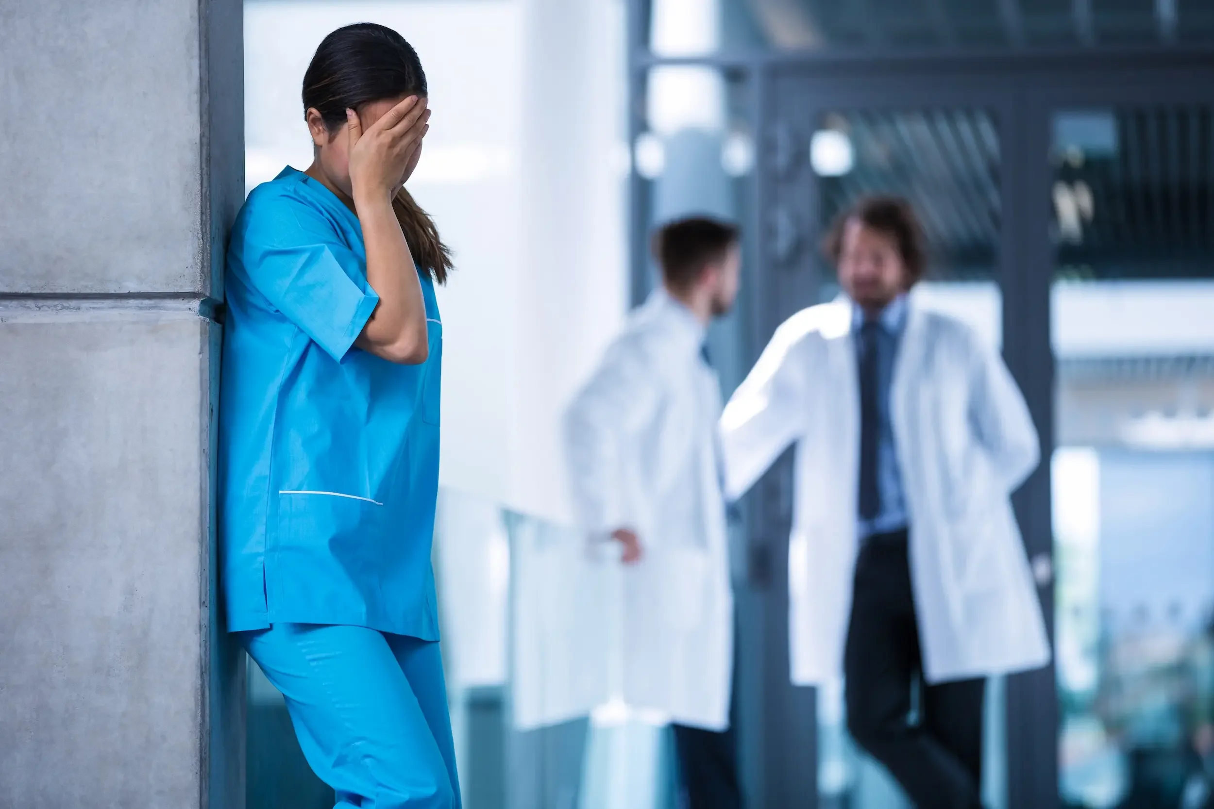 A healthcare worker in scrubs leans against a wall covering her face, embodying physician stress, while two frustrated doctors in lab coats talk in the background.
