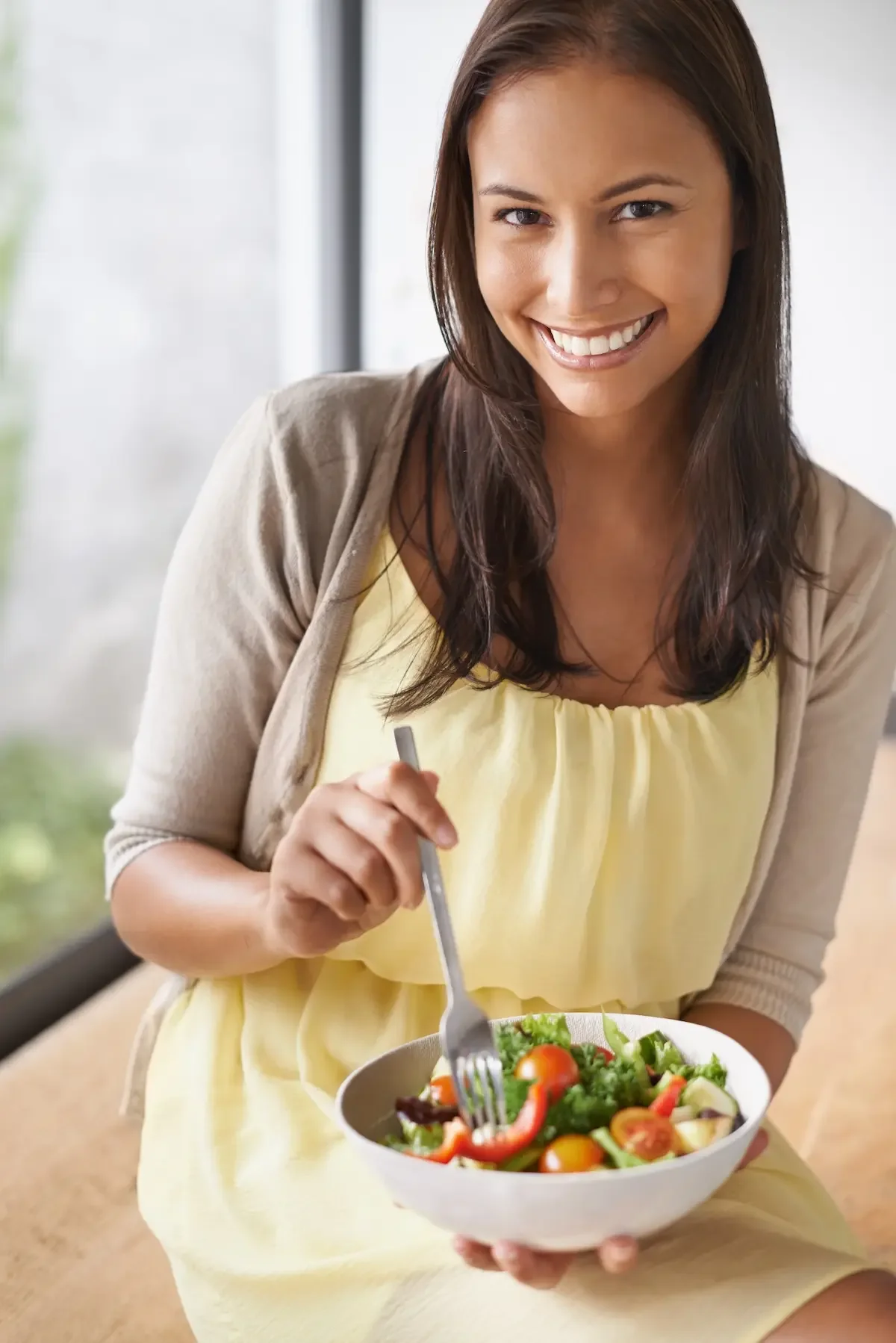 A woman in a yellow dress sits indoors, smiling at the camera while enjoying healthy eating—holding a fork and a bowl of salad filled with various vegetables.