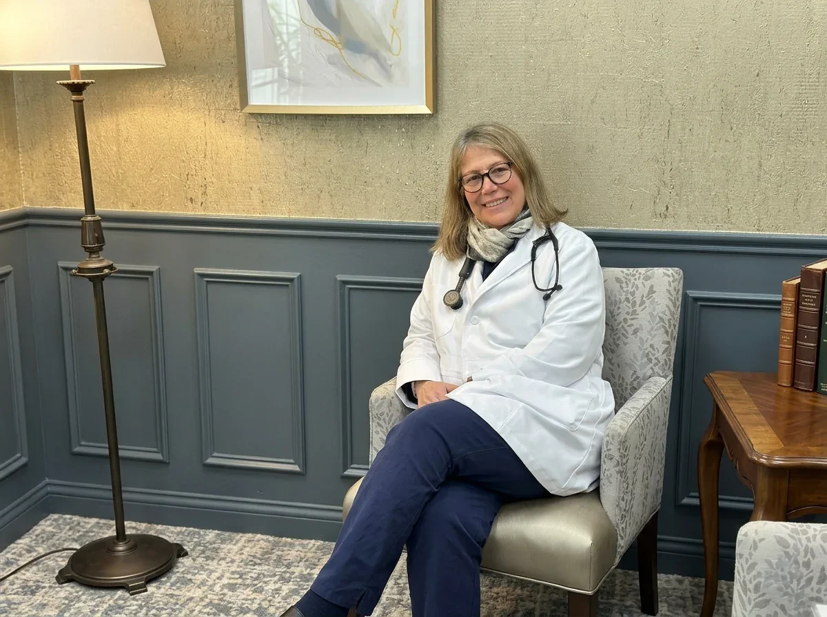 Dr. Anna Manatis, MD, internal medicine physician on Cape Cod, MA, seated in a warm office setting wearing a white coat and stethoscope