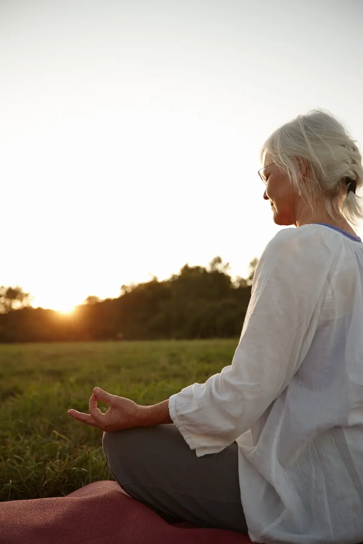 A midlife woman with gray hair sits cross-legged on a mat outdoors at sunset, practicing mindfulness with one hand in a mudra gesture, surrounded by grass and trees.