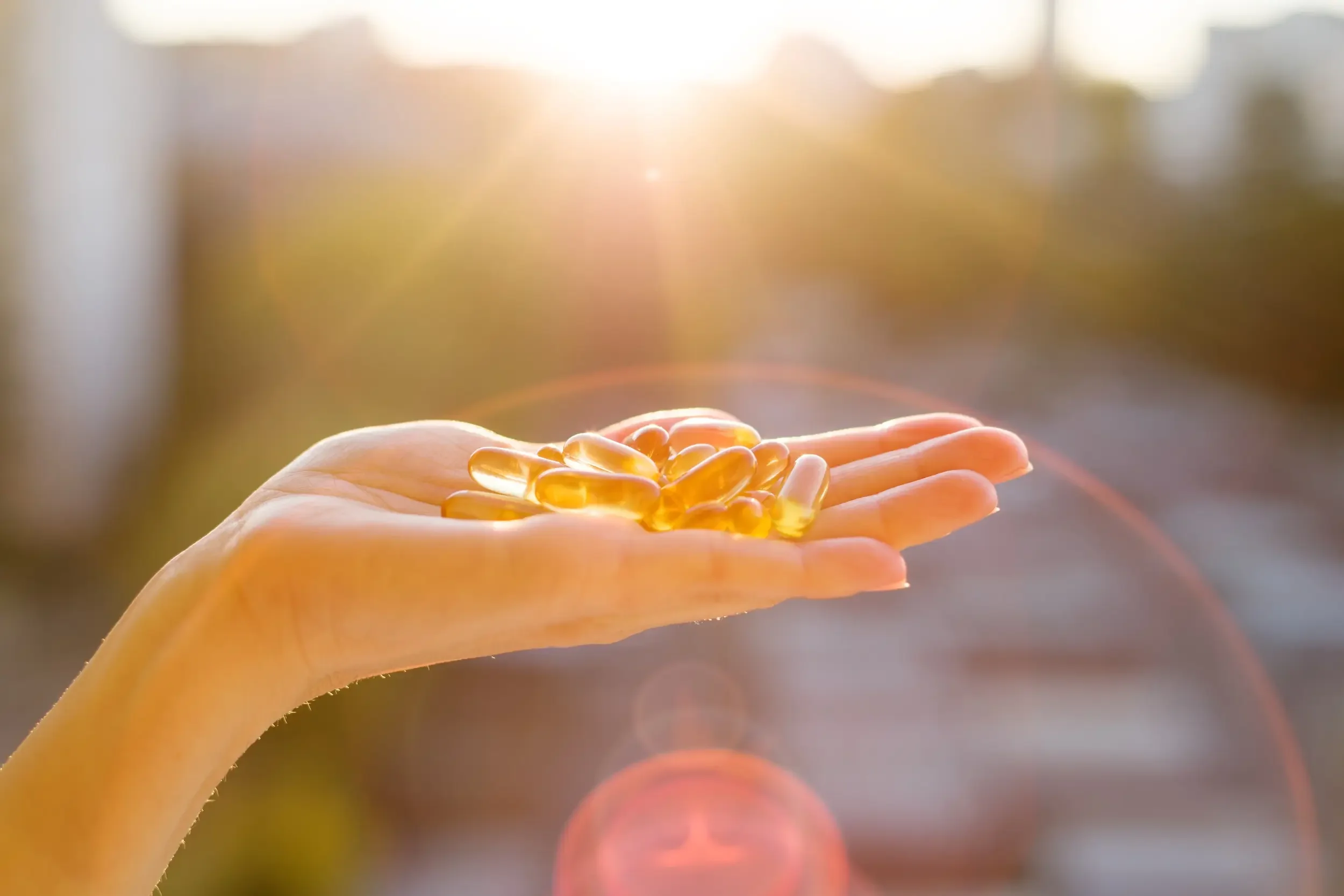 A hand holds several yellow vitamin D softgel capsules outdoors with sunlight and lens flare in the background, highlighting vitamin D benefits.