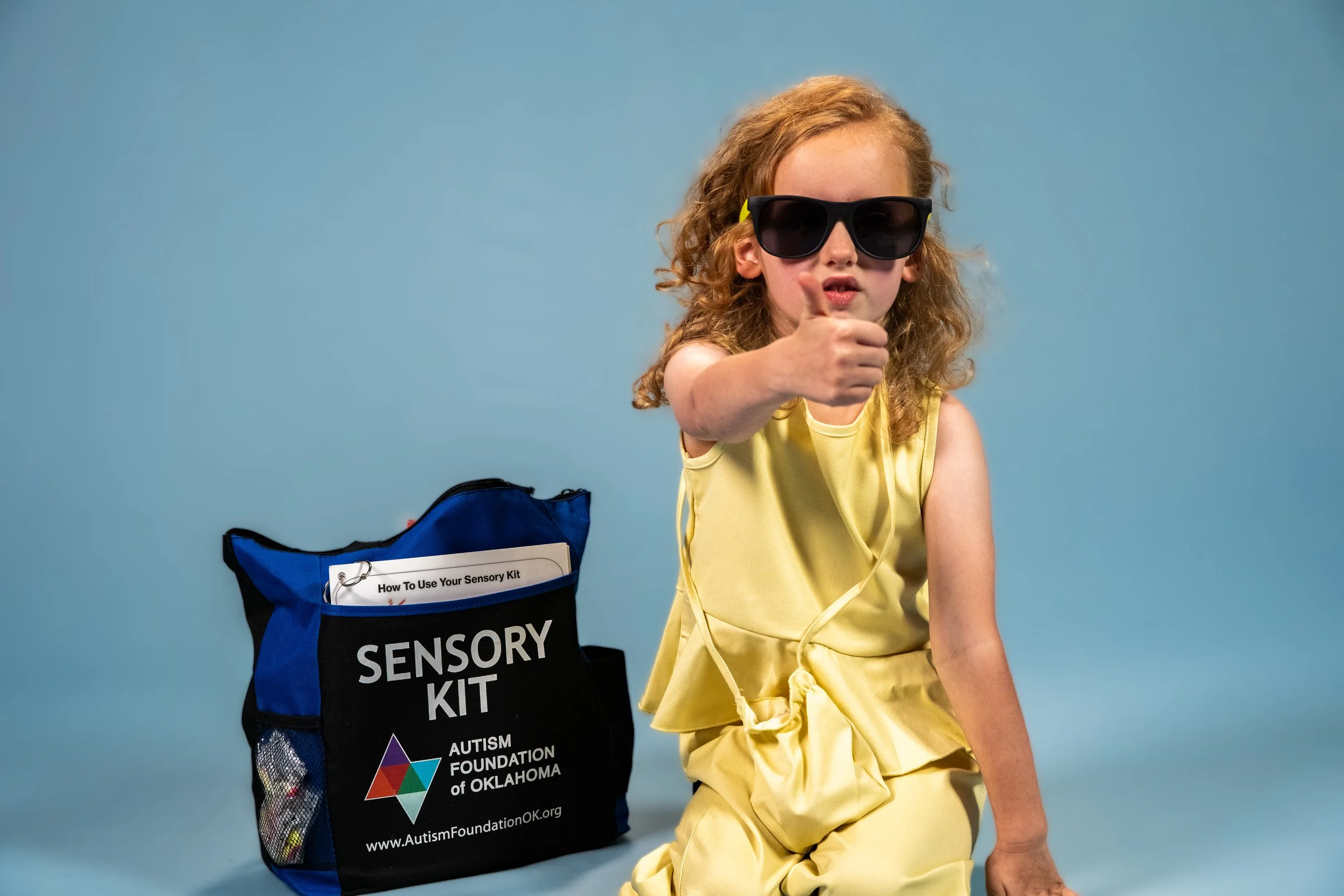 A small female child with sunglasses on, giving a thumbs upsitting next to an AFO sensory kit