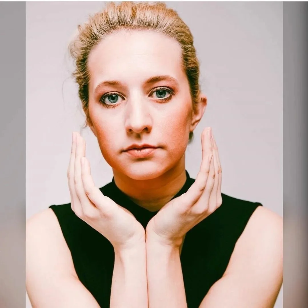 Porträt einer nachdenklichen Frau mit hellem Haar in einem schwarzen Oberteil, die ihre Hände vor ihrem Gesicht hält.