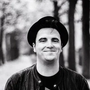 Schwarz-weiß Porträt eines lächelnden Mannes mit Hut und Lederjacke, unscharfer Hintergrund mit Bäumen.