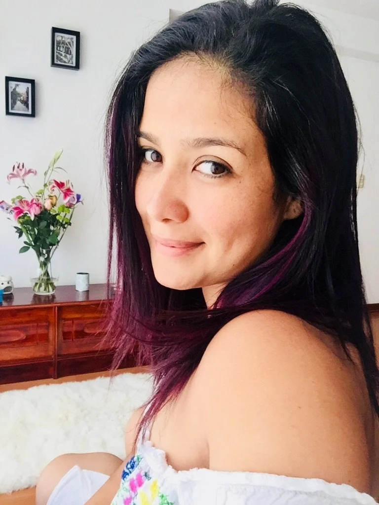 Retrato de una mujer con cabello negro y con puntas violetas, sonriendo ligeramente, en un ambiente interior con flores y cuadros en la pared.