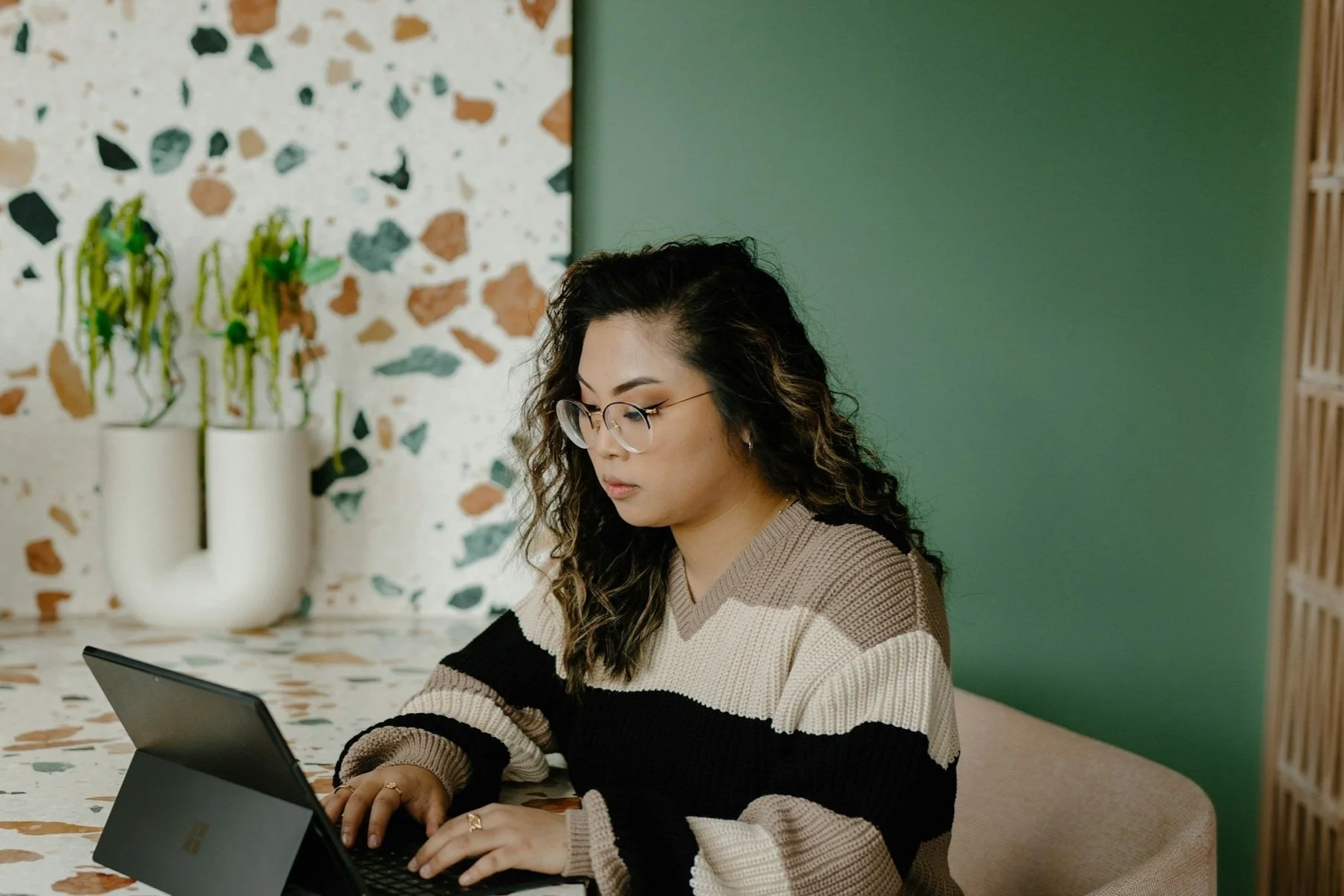 A young woman with curly black hair, glasses, and a beige and black striped sweater working on a tablet at a terrazzo table.