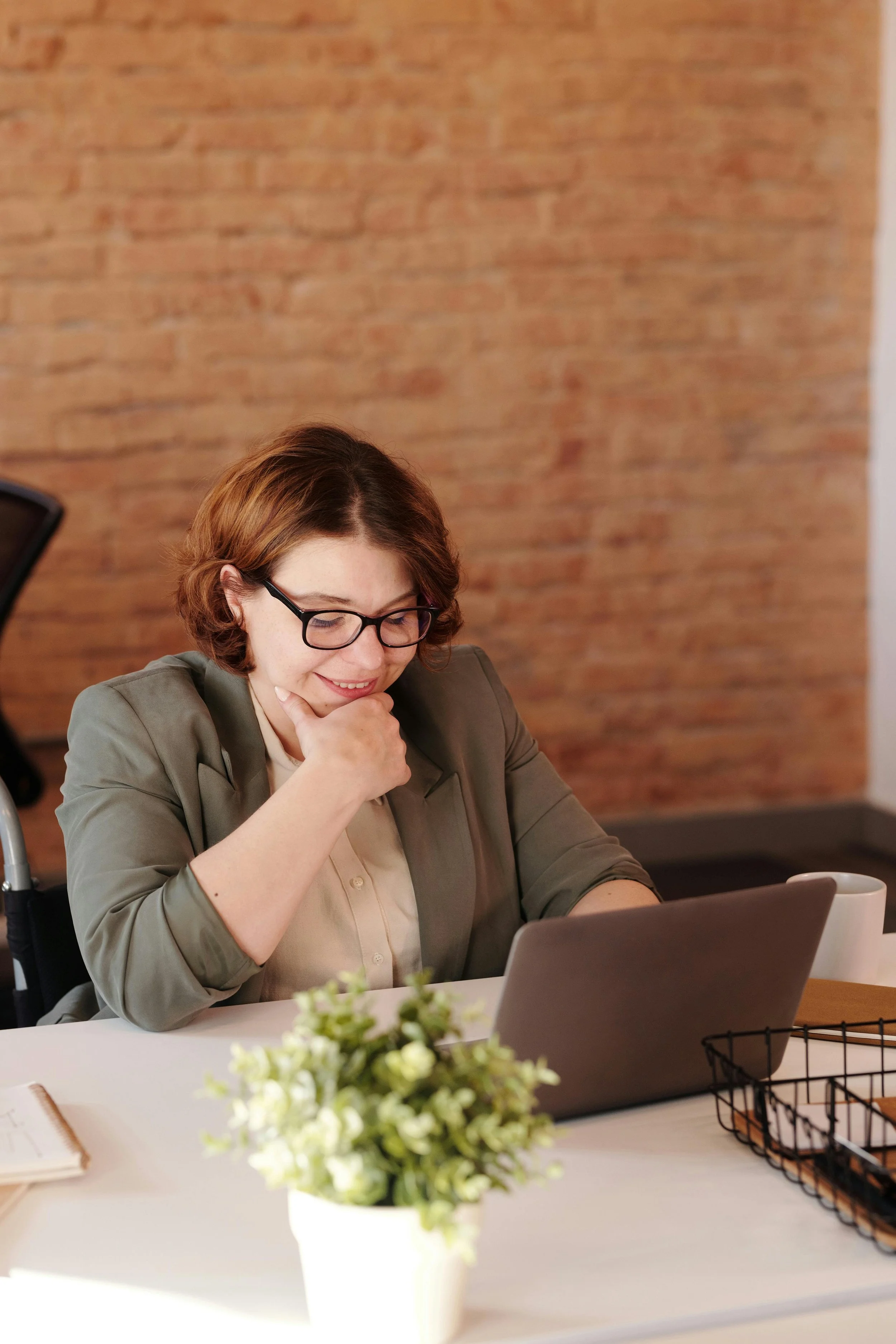 A woman with short brown hair, glasses, and a beige shirt under a green blazer sitting at a desk looking at a laptop, with a small plant, a notebook, and a wire basket on the desk, and a brick wall in the background.