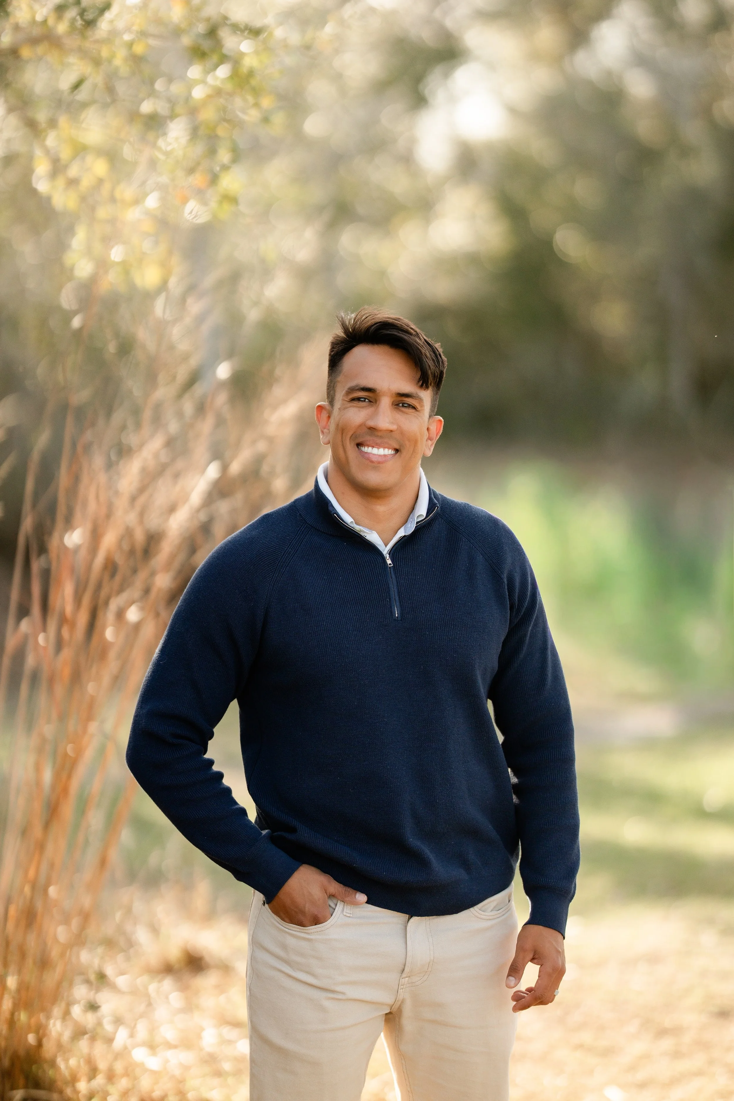 A young man smiling outdoors wearing a navy blue sweater and light-colored pants, standing on a nature trail with trees and foliage in the background.