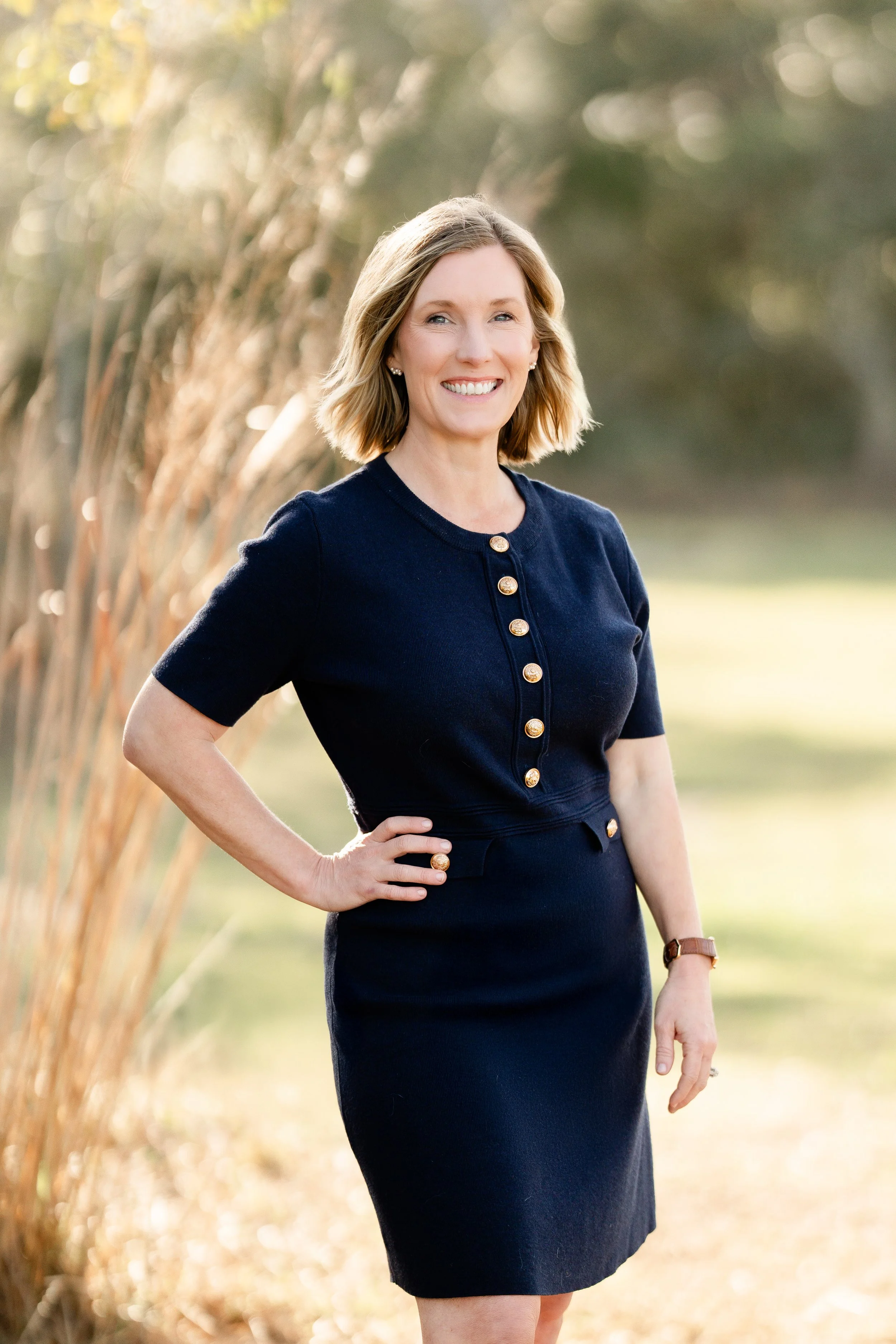 A woman smiling outdoors, wearing a navy blue dress with gold buttons.