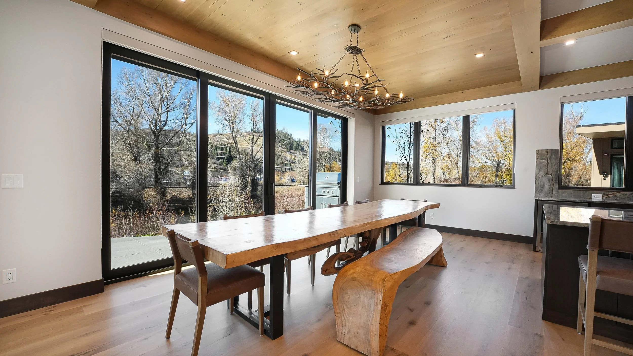 Rustic-modern dining room with live-edge wood table, branch chandelier, timber ceiling, and sliding glass doors with Colorado hillside views — Refine Imagery listing photography