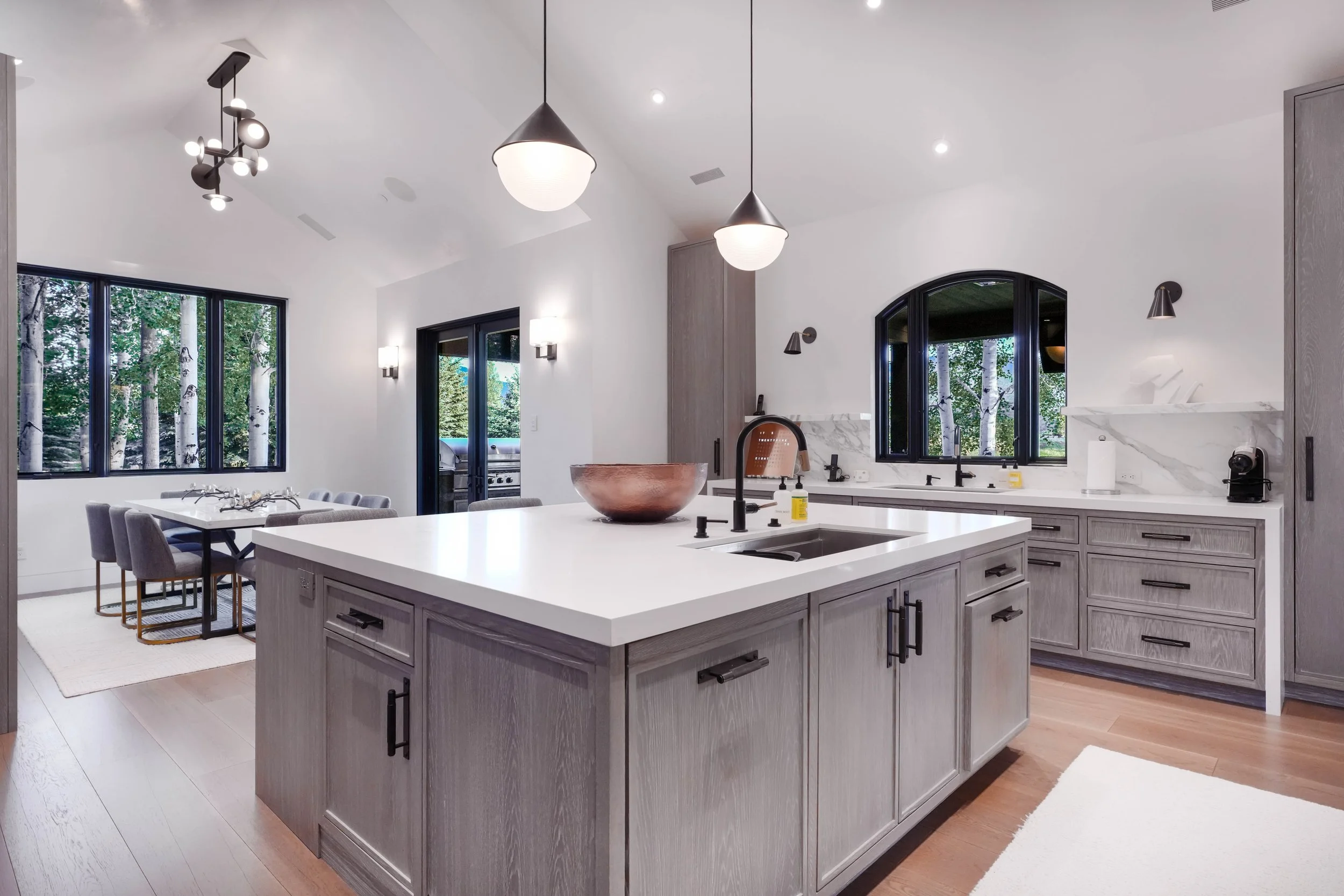 Open-concept luxury kitchen with gray oak cabinetry, quartz island countertop, pendant lighting, and aspen tree views through black-framed windows — professional real estate photography by Refine Imagery