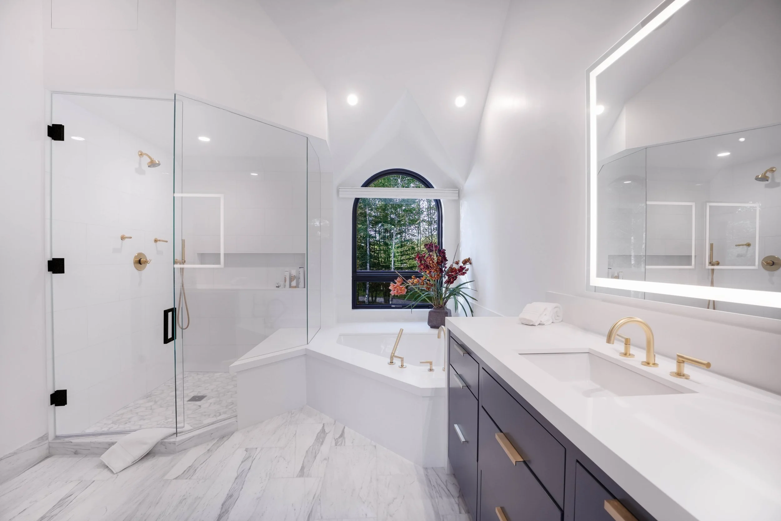 Luxury bathroom with glass walk-in shower, corner soaking tub, brushed gold fixtures, backlit vanity mirror, and arched window — real estate interior photography by Refine Imagery