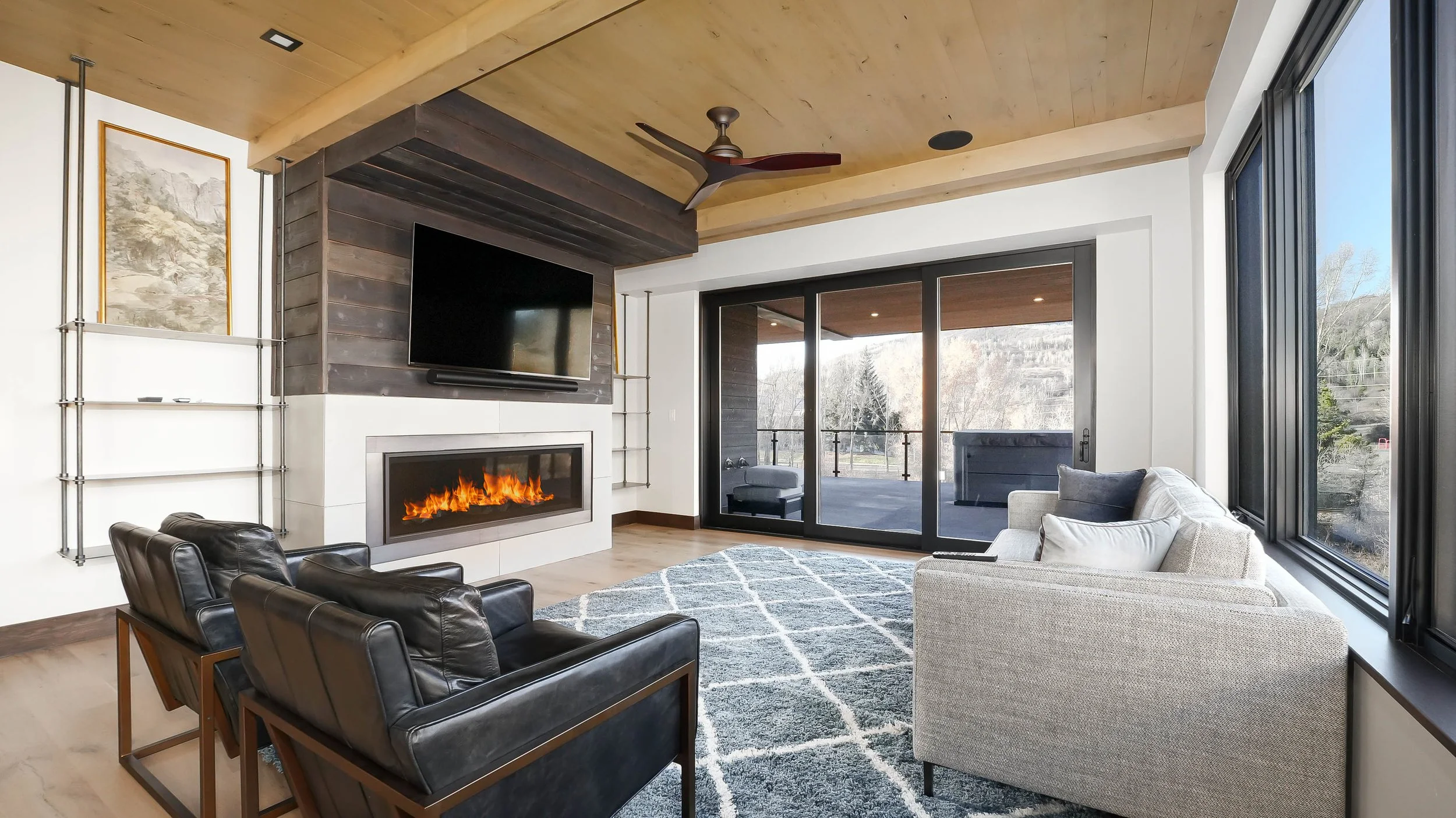 Modern mountain living room with linear gas fireplace, dark wood accent wall, leather chairs, timber ceiling, and sliding doors to balcony with hillside views — Colorado luxury home photography by Refine Imagery