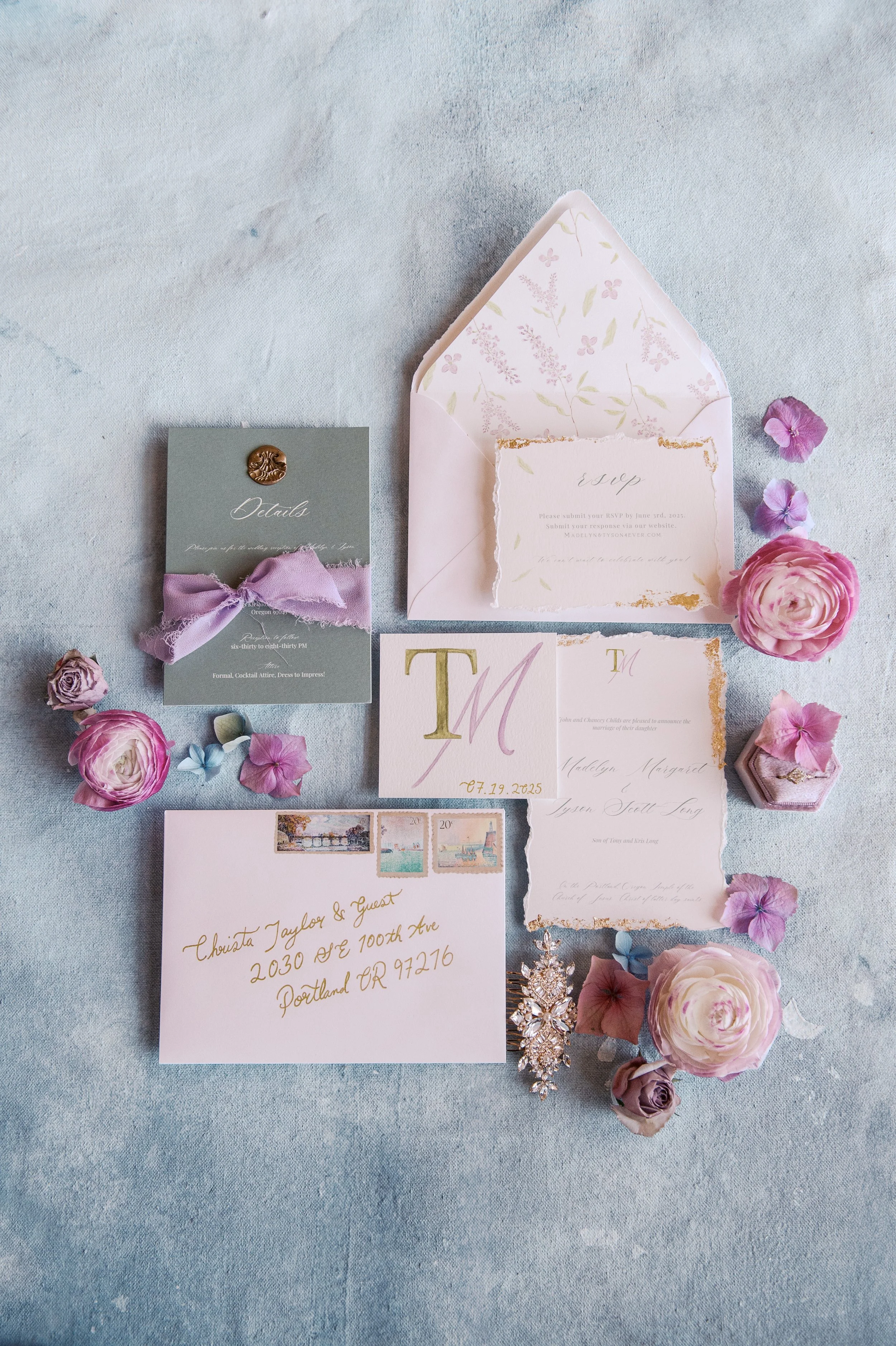 Wedding invitation suite with pink flowers, RSVP card, envelope, guest address card, and jewelry, arranged on light blue fabric.