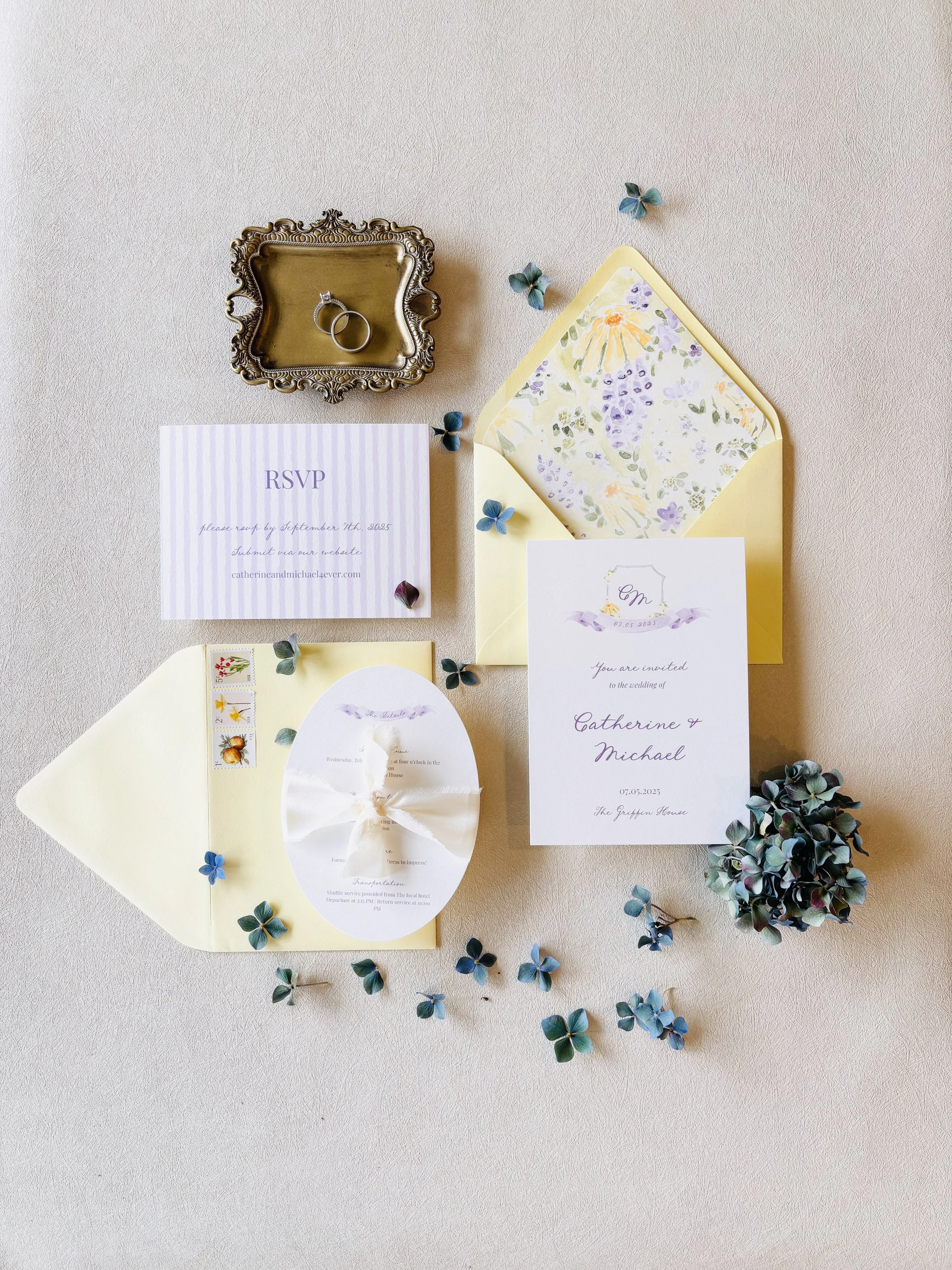 Wedding invitation suite with RSVP card, envelope, and decorative blue and purple flowers, featuring rings, a small silver tray, and a sprig of hydrangea on a light background.
