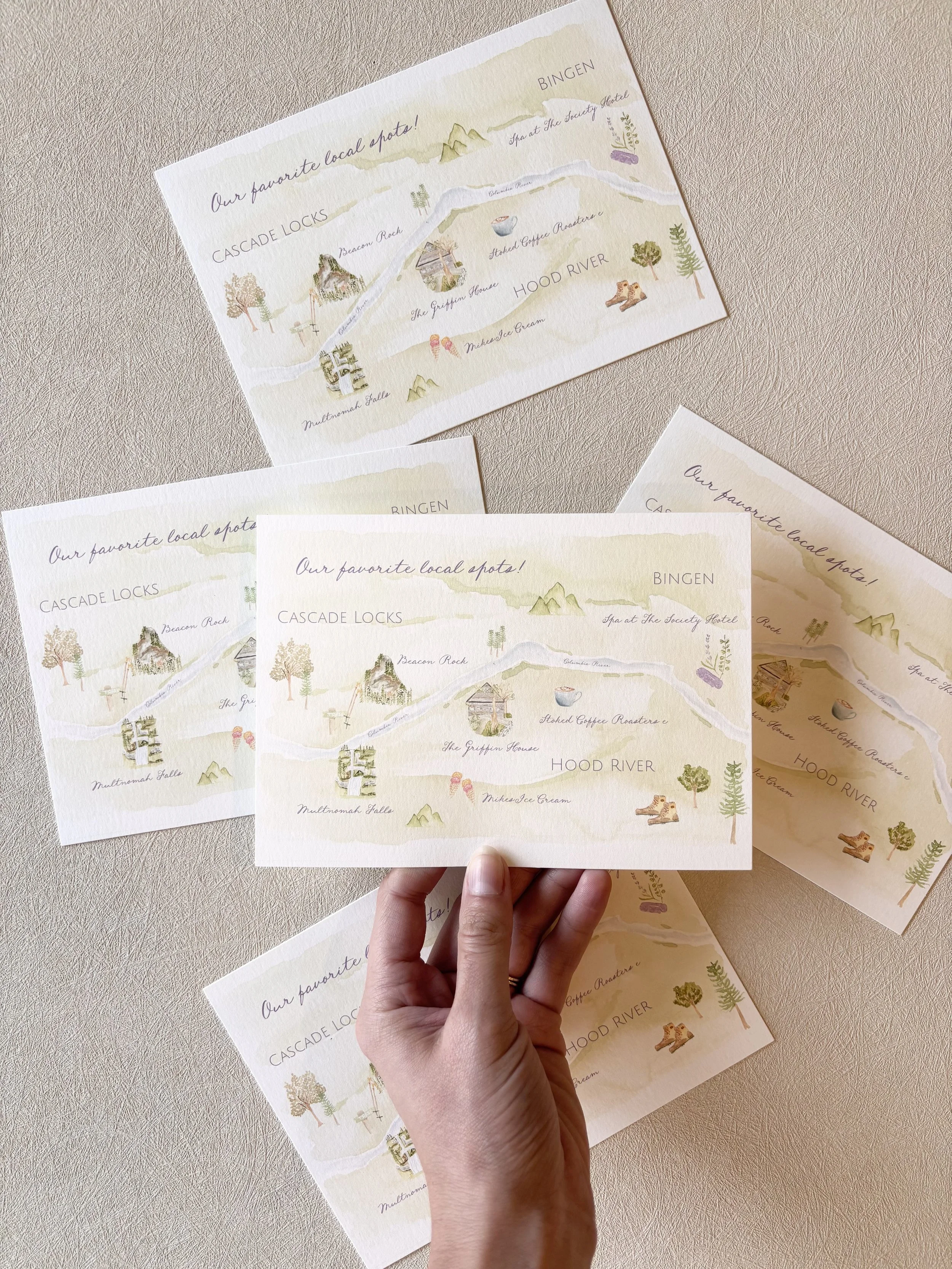 A hand holding a illustrated map of a local area featuring Cascade Locks, Bingen, and Hood River, with landmarks like Beacon Rock, The Griffin House, and Multnomah Falls, along with decorative trees and mountains.
