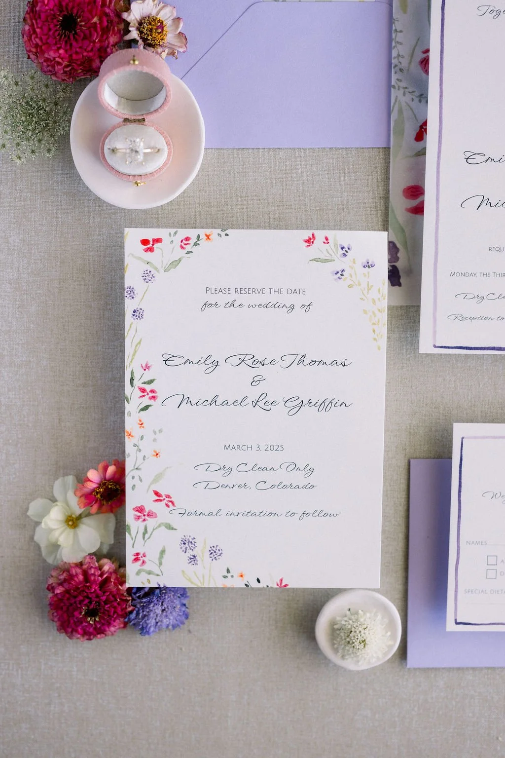 Wedding invitation with floral border, surrounded by flowers and a pink ring box with wedding rings.