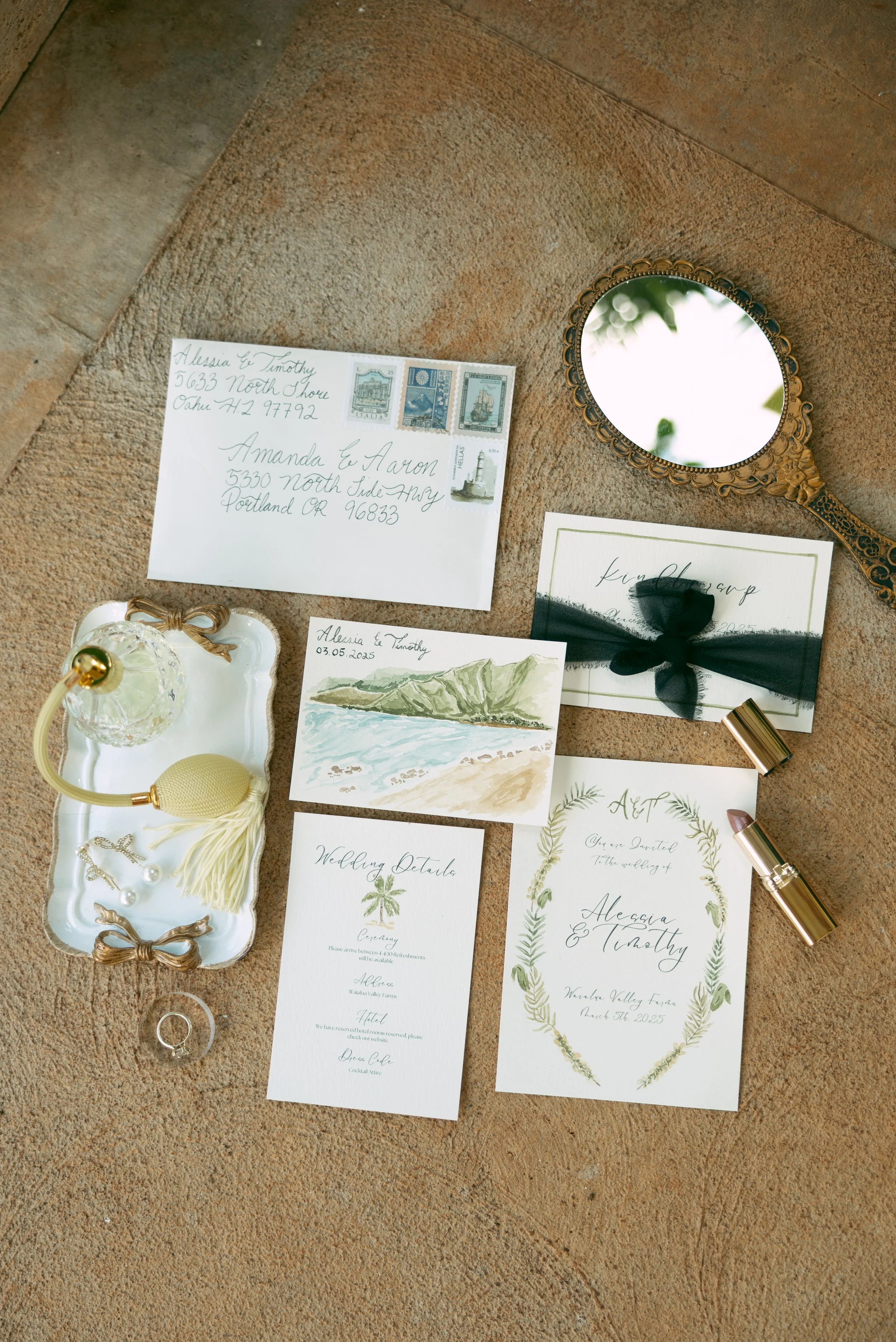 Wedding invitation suite with RSVP card, watercolor coast scene, palm tree wedding details, and an address envelope, all arranged on a textured surface. Additional items include a miniature mirror, perfume, and lipstick.