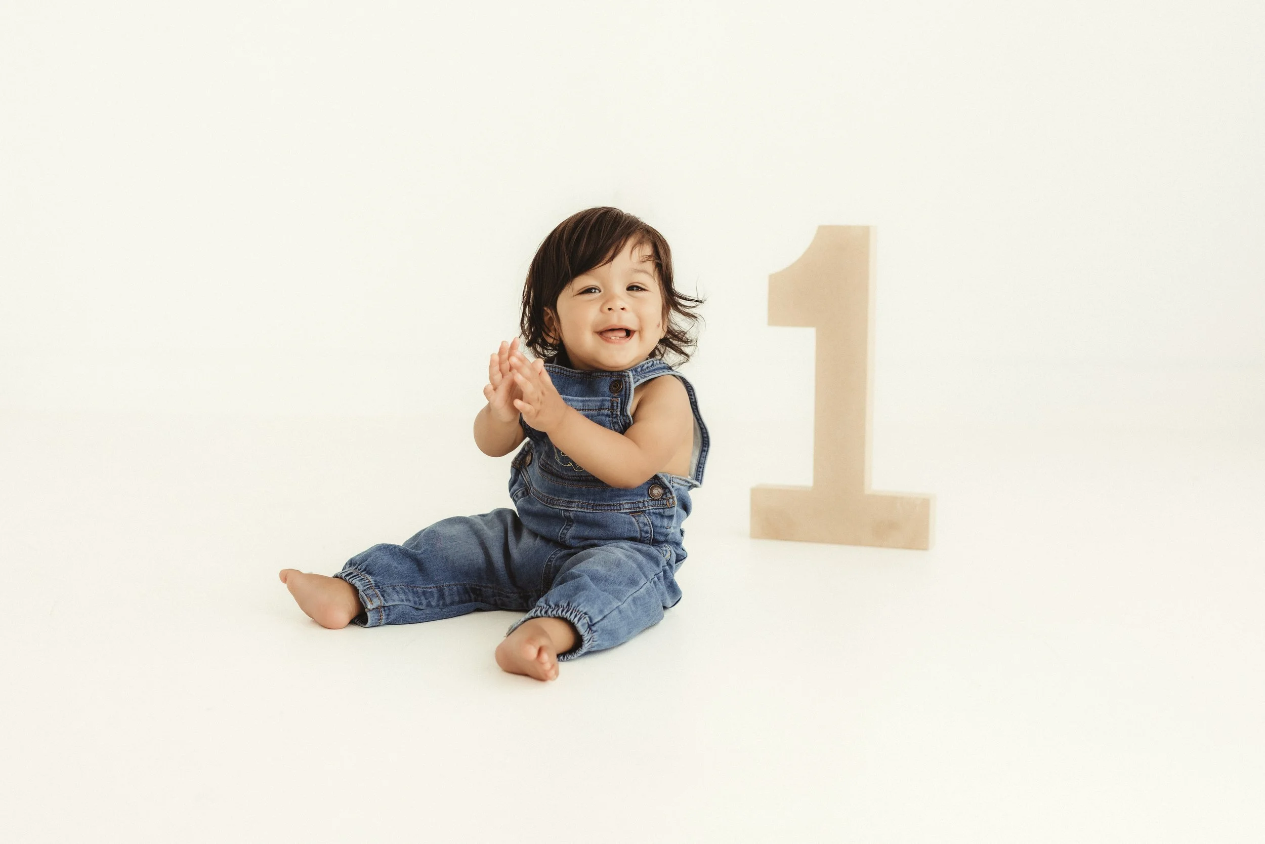 A smiling young child with dark hair, wearing denim overalls, sitting barefoot on a white surface, clapping hands, and a large beige wooden number one standing upright behind them, set against a plain white background.