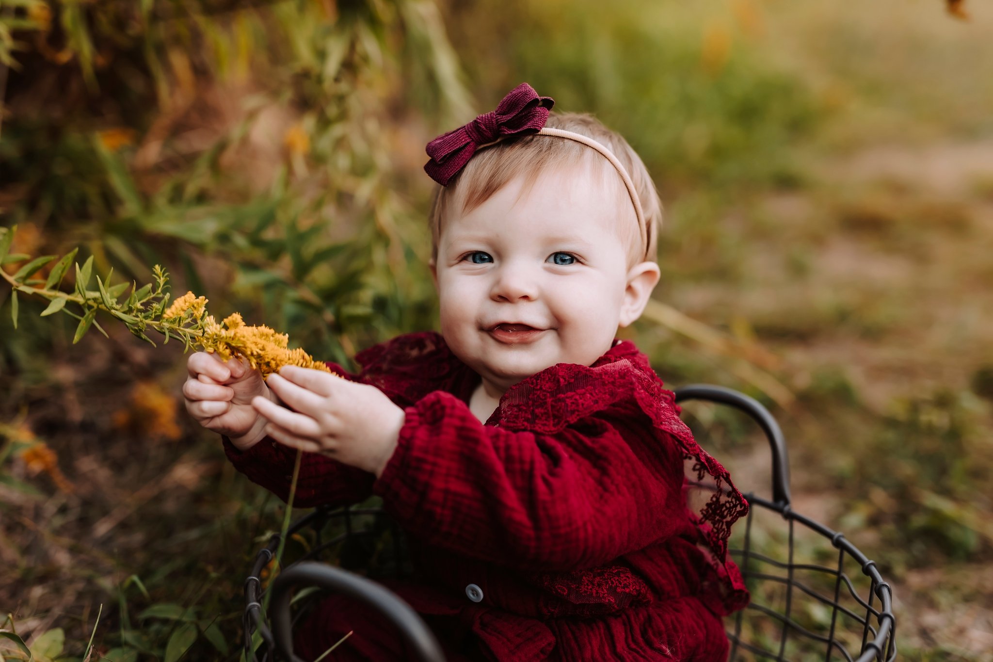A smiling young child with blonde hair and blue eyes, wearing a red dress and a headband with a bow, sitting in a stroller outdoors among autumn foliage, holding a yellow flower.