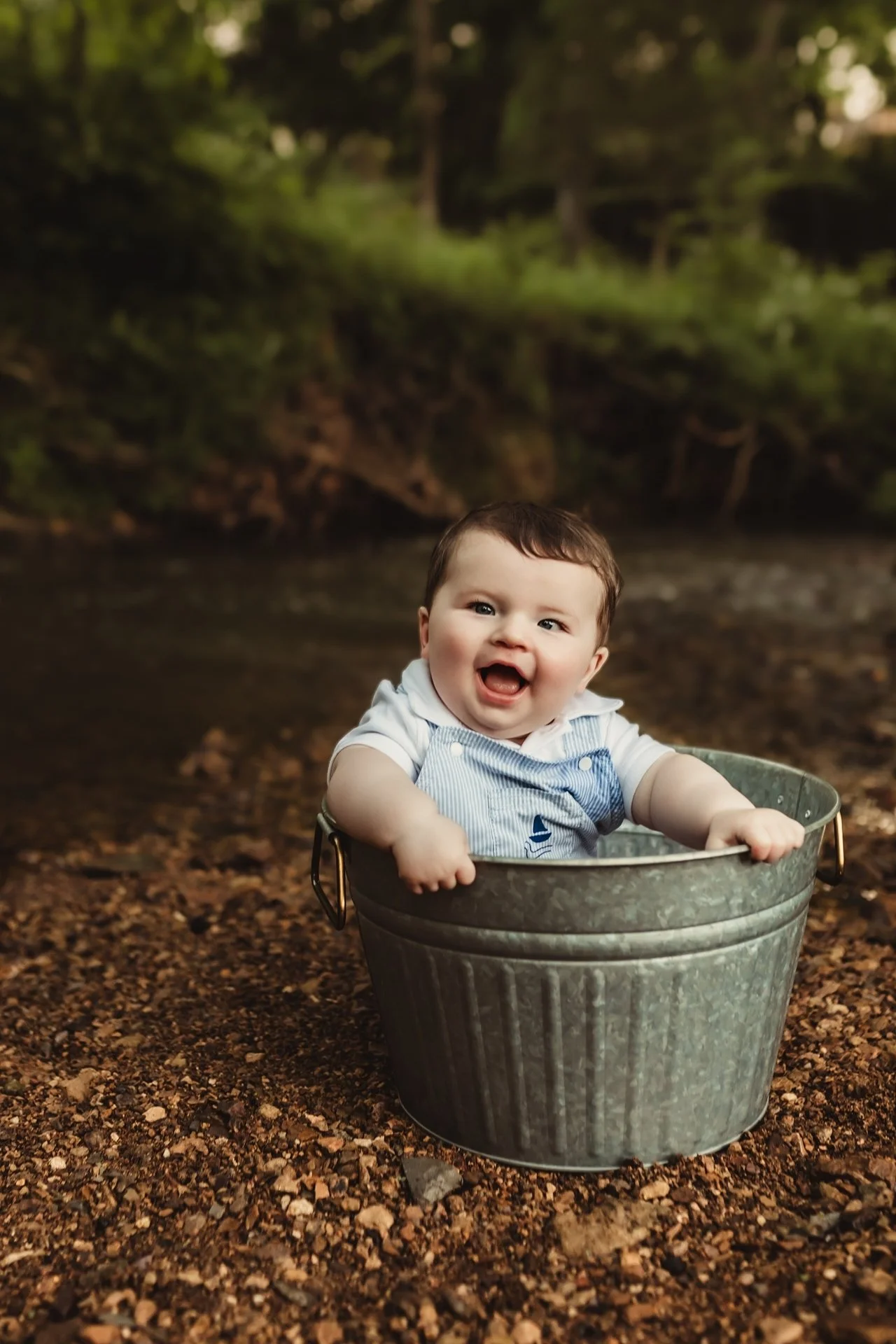 A happy baby sitting in a metal bucket outdoors on a ground covered with small rocks and dirt, with a background of trees and foliage.