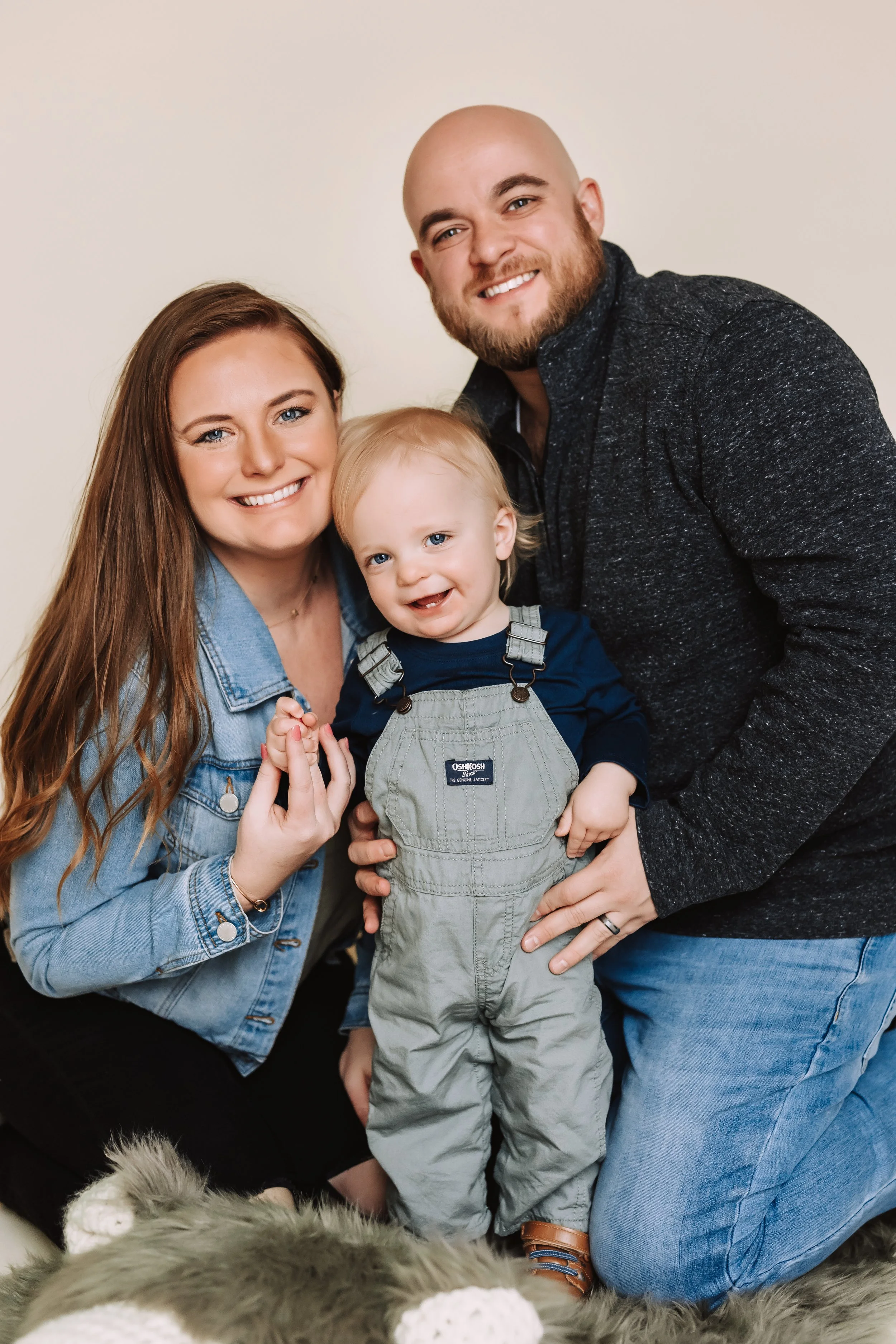A happy family of three, mom, dad, and a young boy, smiling and posing together indoors with a plain background. They are close together; the mom and dad are on either side of the boy, who is in the center. The mother has long brown hair, the father 
