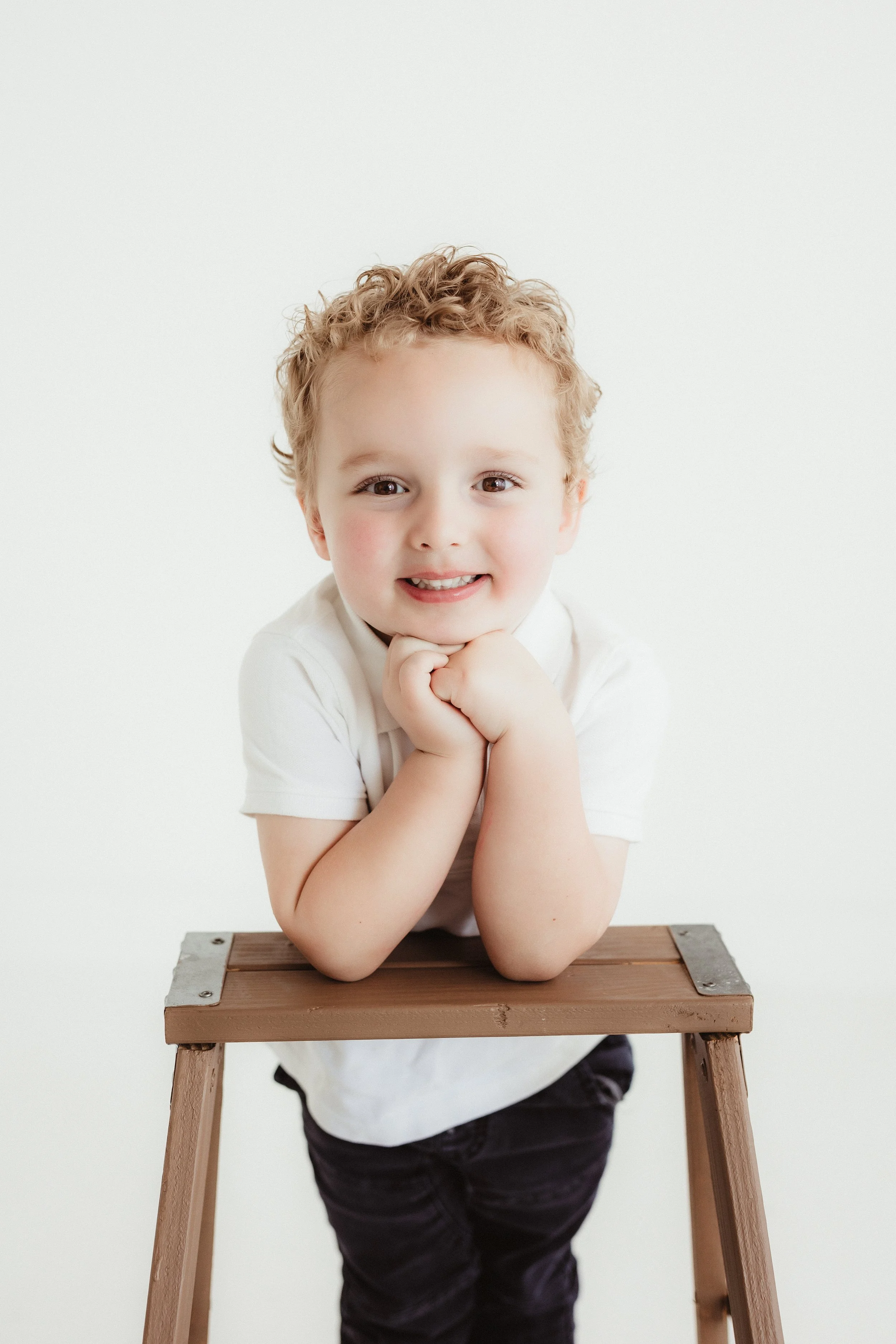 A young boy with curly blonde hair leaning on a wooden stool with his chin resting on his hands, smiling at the camera against a plain white background.