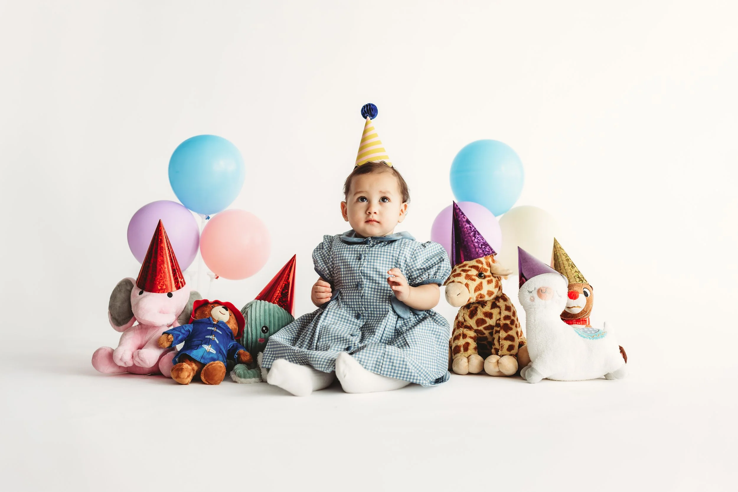 A young child sitting surrounded by colorful balloons and plush toys on a plain white background, wearing a party hat and a checkered dress.