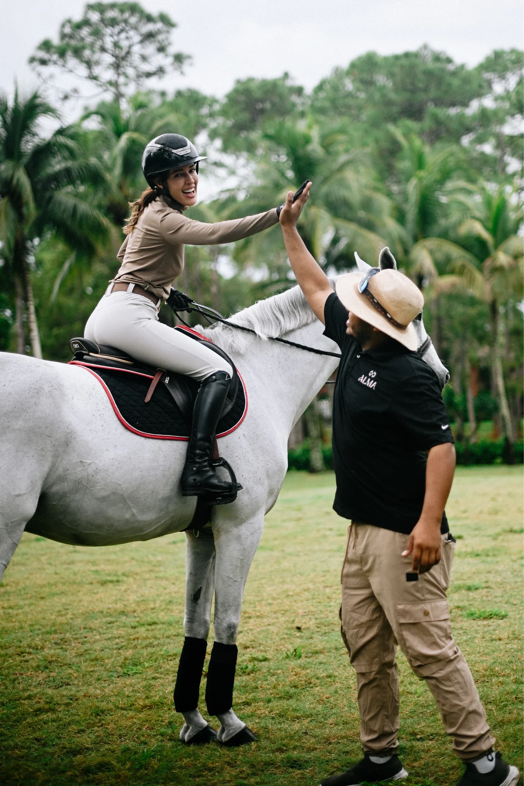 A woman riding a white horse and smiling, wearing a beige riding outfit and helmet, giving a high-five to a man standing next to the horse, who is wearing a wide-brimmed hat, black shirt, and tan pants, in a lush, green outdoor setting with trees in 