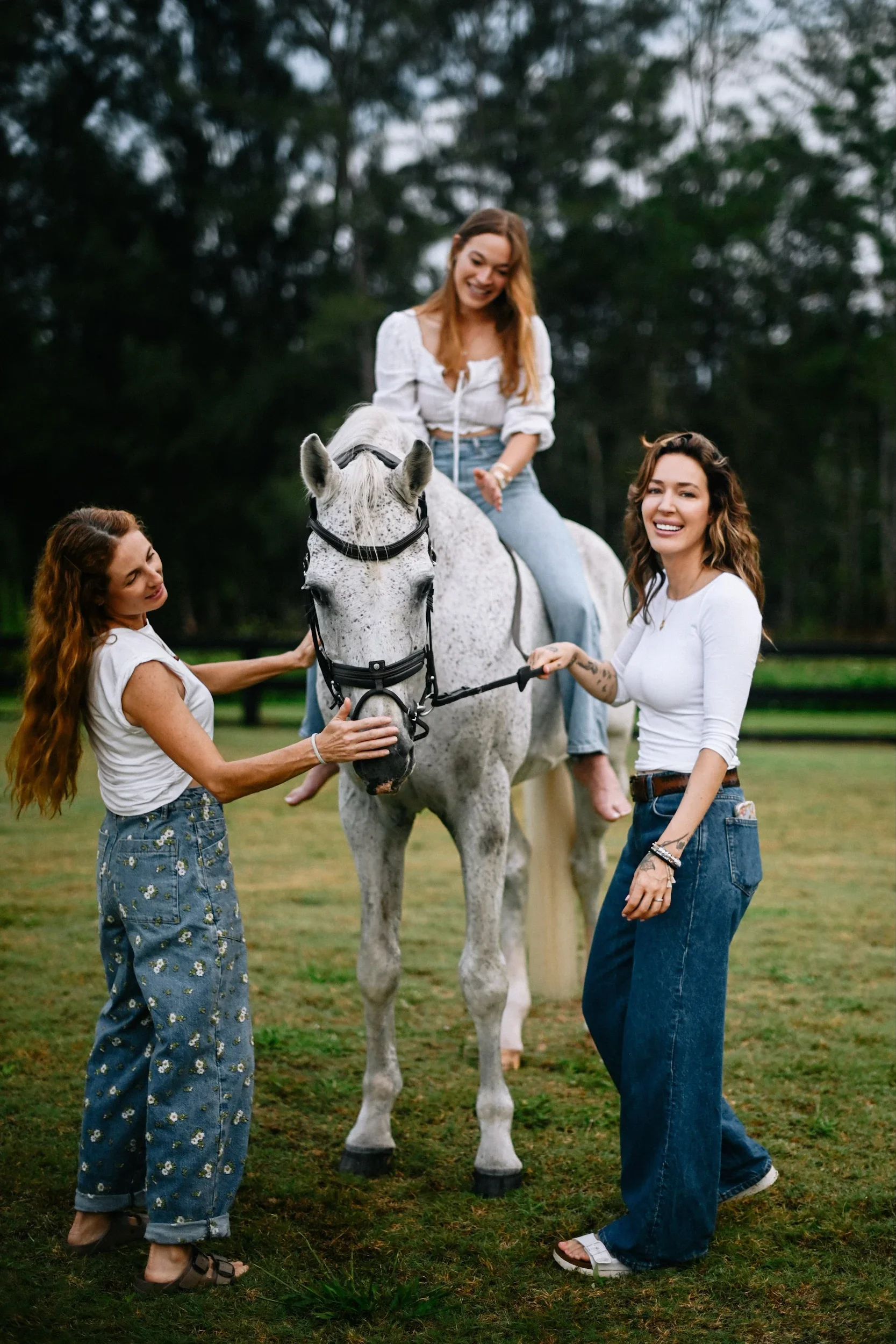 Three women and a white horse on a grassy field, with the woman on the horse smiling and two women holding the horse's reins.