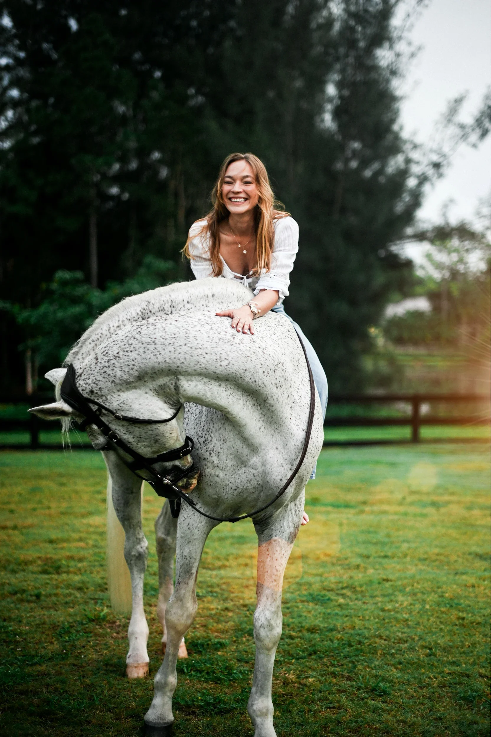 A woman with long, wavy, light brown hair smiling while riding a white and gray-speckled horse in a grassy, wooded area.